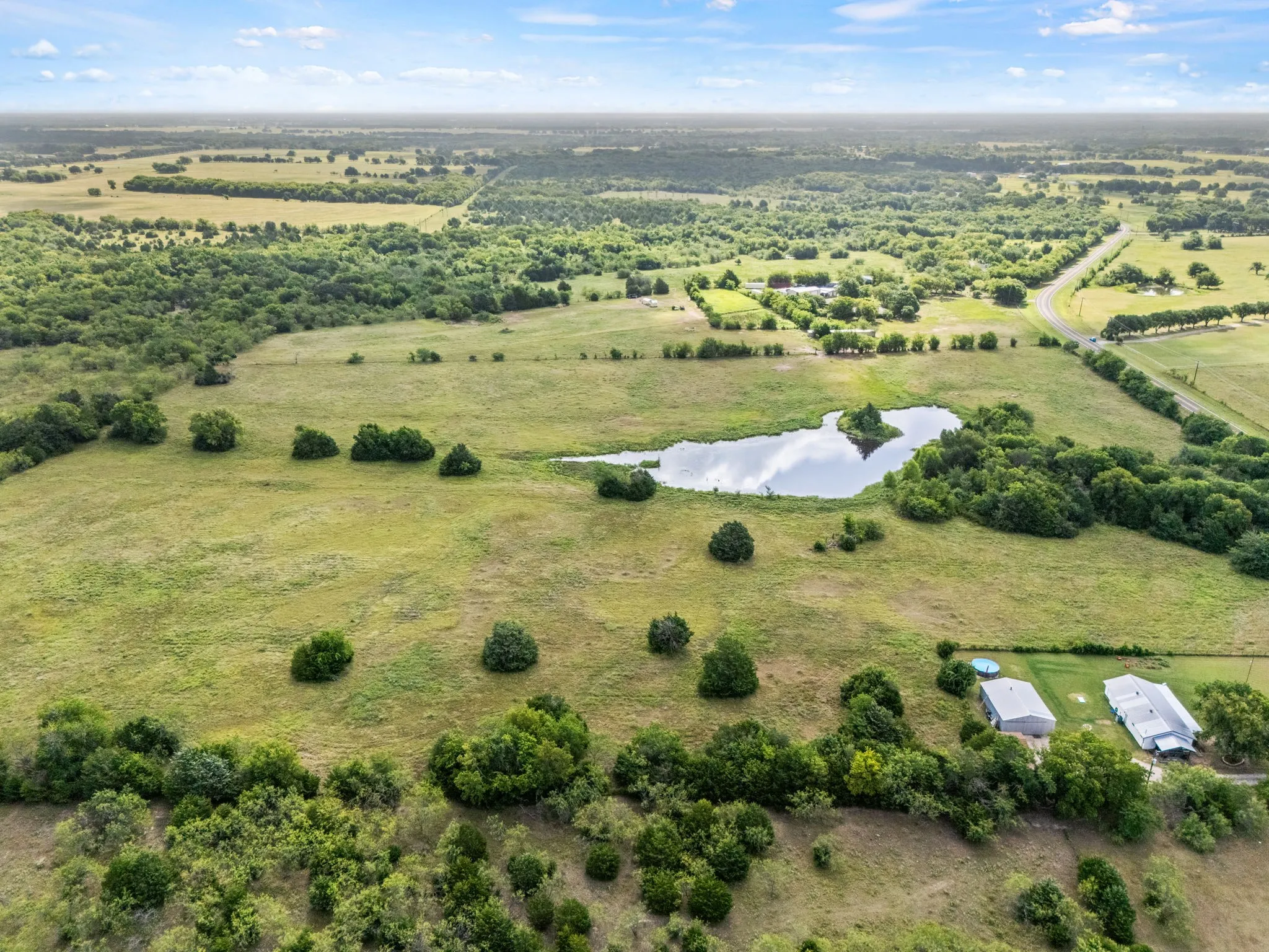 Expansive land parcel featuring a natural pond, mature trees, and open grassy areas, with two single-story structures and a paved road visible in the distance