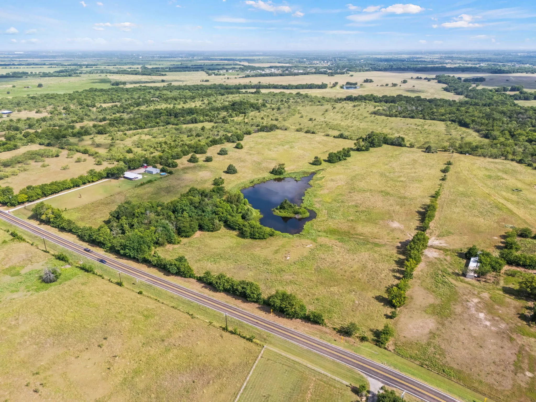 Expansive rural property featuring a private pond with a central island, verdant tree lines, and a paved road frontage