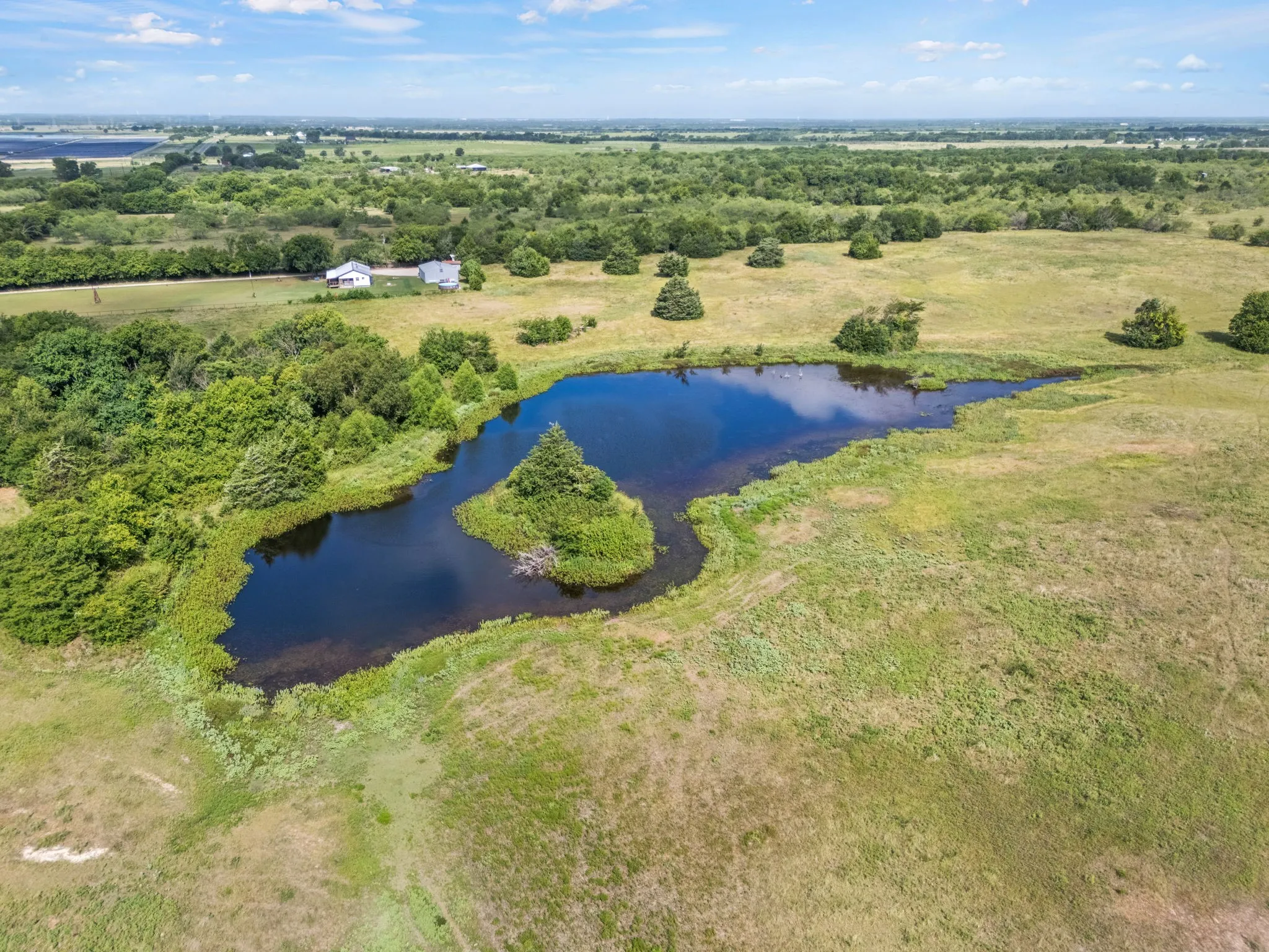 Expansive rural landscape featuring a prominent pond with an island, surrounded by natural grasslands and mature trees