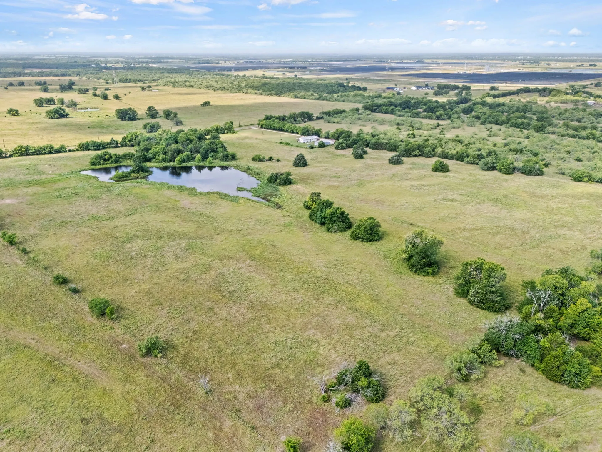 Expansive rural landscape featuring a natural pond surrounded by varied green foliage, open fields, and scattered trees under a partially cloudy sky