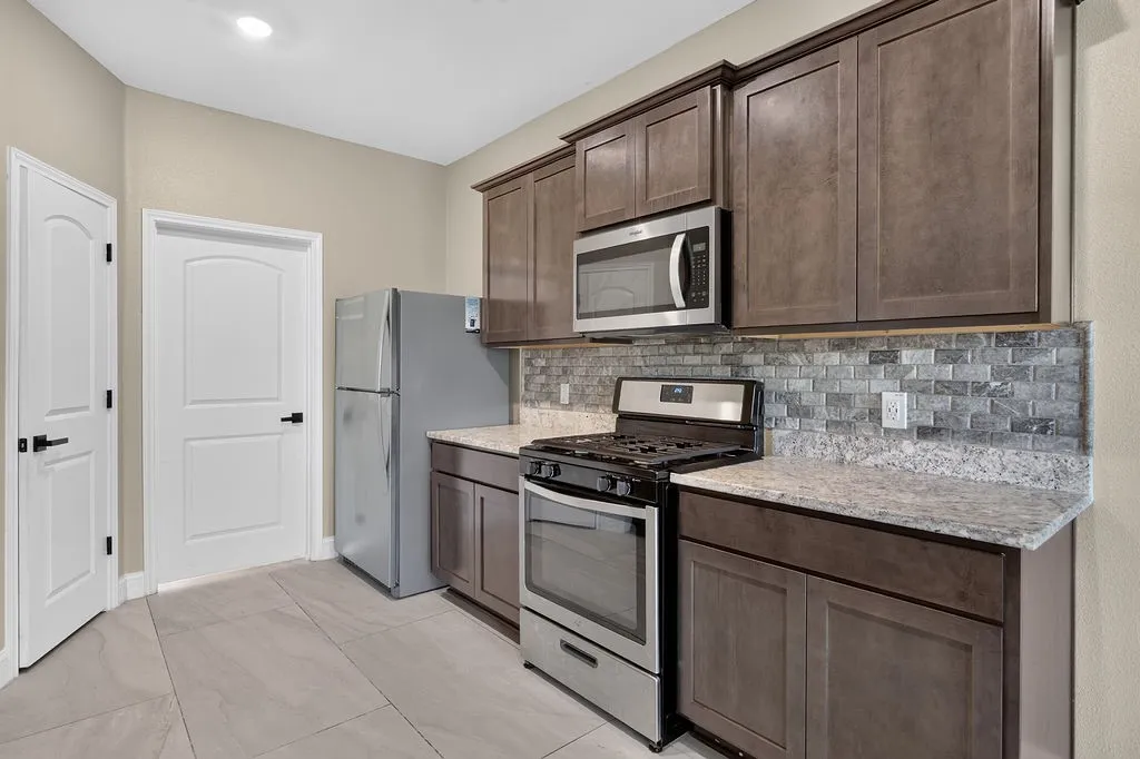 Kitchen featuring wood-finish cabinetry, light-toned granite countertops, a subway tile backsplash, and stainless steel appliances