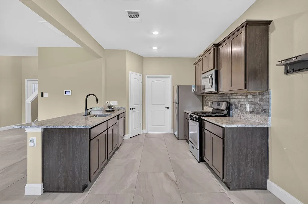 Open-concept kitchen featuring granite countertops, dark wood-finish cabinetry, stainless steel appliances, a tile backsplash, and light-toned tile flooring