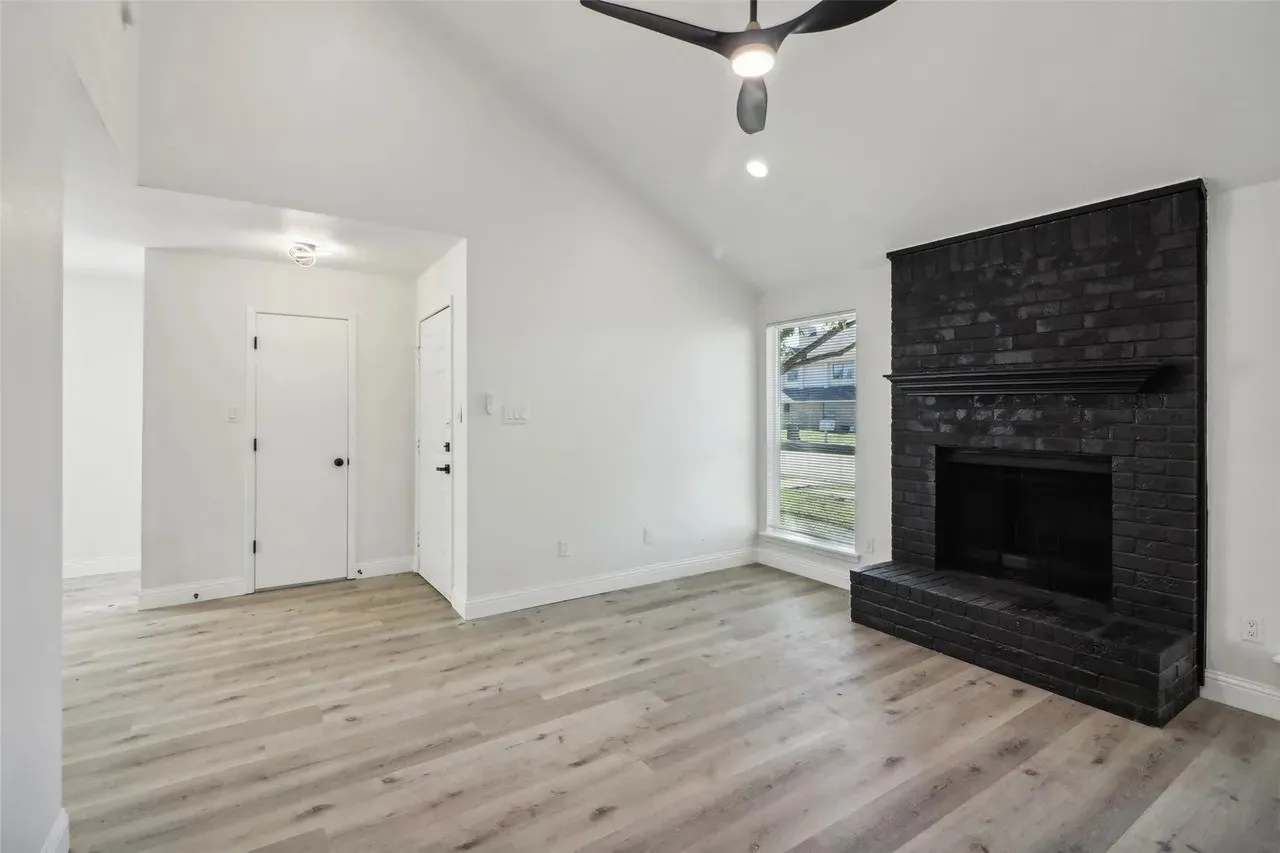 Vaulted ceiling living area featuring a prominent black brick fireplace with mantel, light wood-finish flooring, a modern black ceiling fan, and recessed lighting