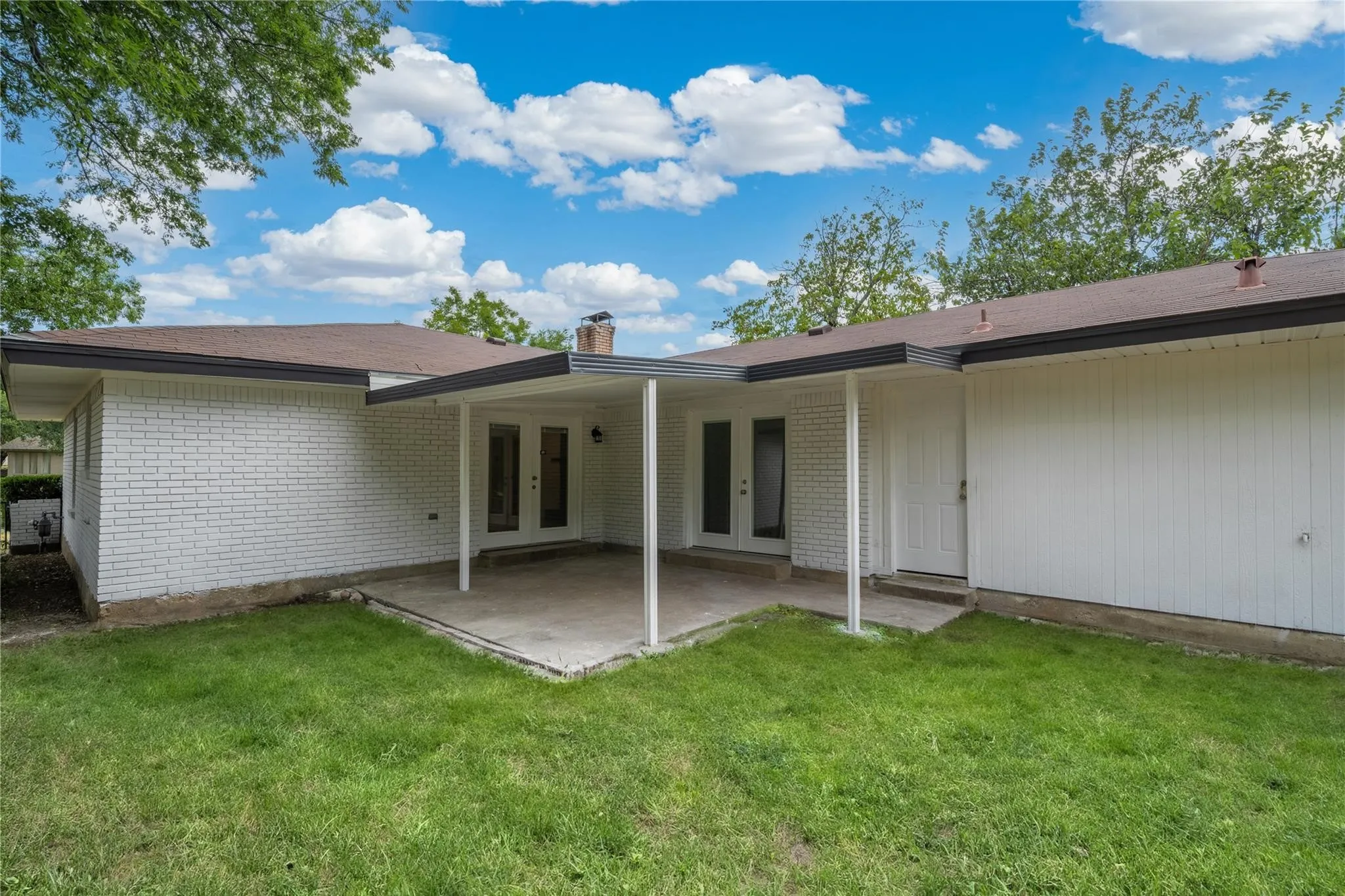Covered patio area with concrete flooring, dual French doors, and an additional exterior door, set against a backdrop of white brick and vertical siding
