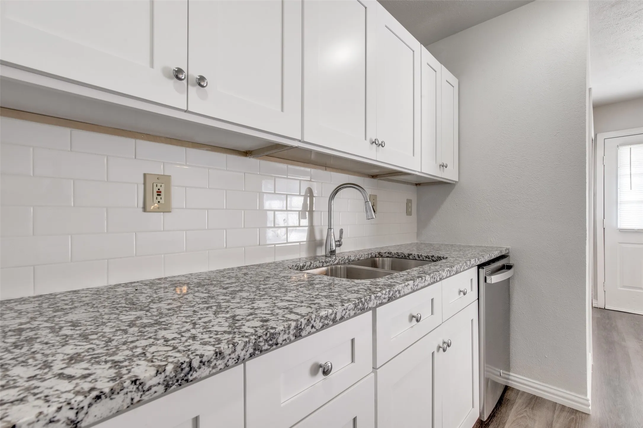 Kitchen featuring white shaker-style cabinetry, a speckled countertop, a double basin stainless steel sink with a high-arc faucet, a white subway tile backsplash, and a built-in dishwasher
