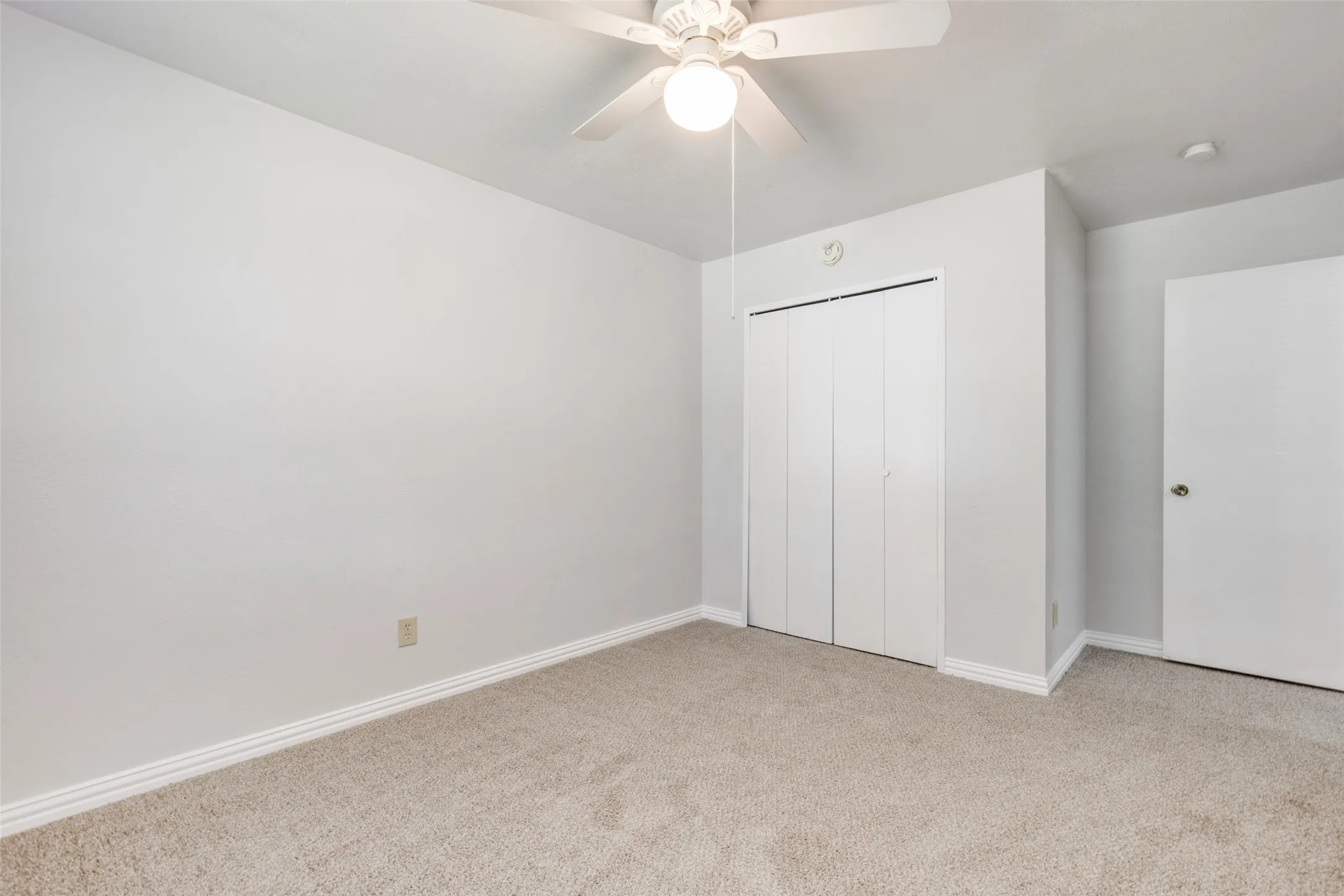 Light-colored carpeting frames a room with a ceiling fan, bi-fold closet doors, and white baseboards