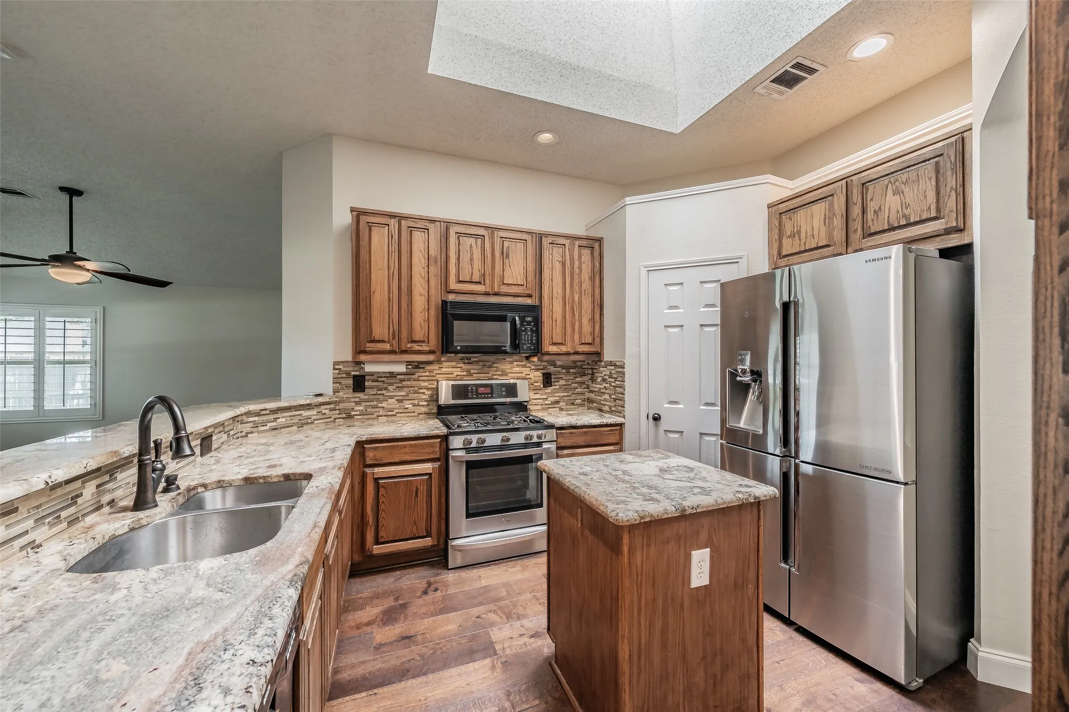 Kitchen featuring wood cabinetry, stainless steel appliances, a stone tile backsplash, a center island with a stone countertop, and wood-finish flooring