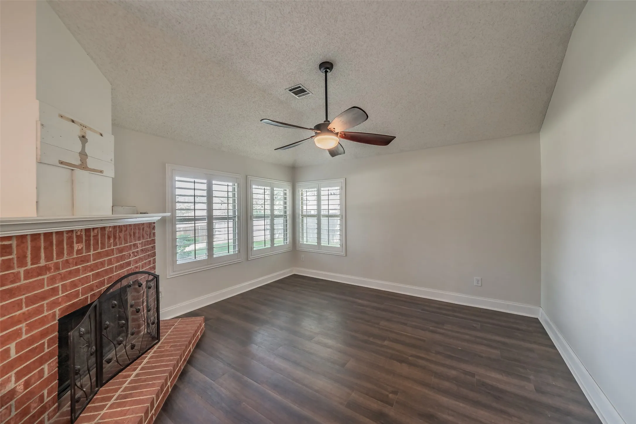 Living area featuring a brick fireplace with a black metal screen, wood-finish flooring, white plantation shutters, and a ceiling fan with integrated lighting