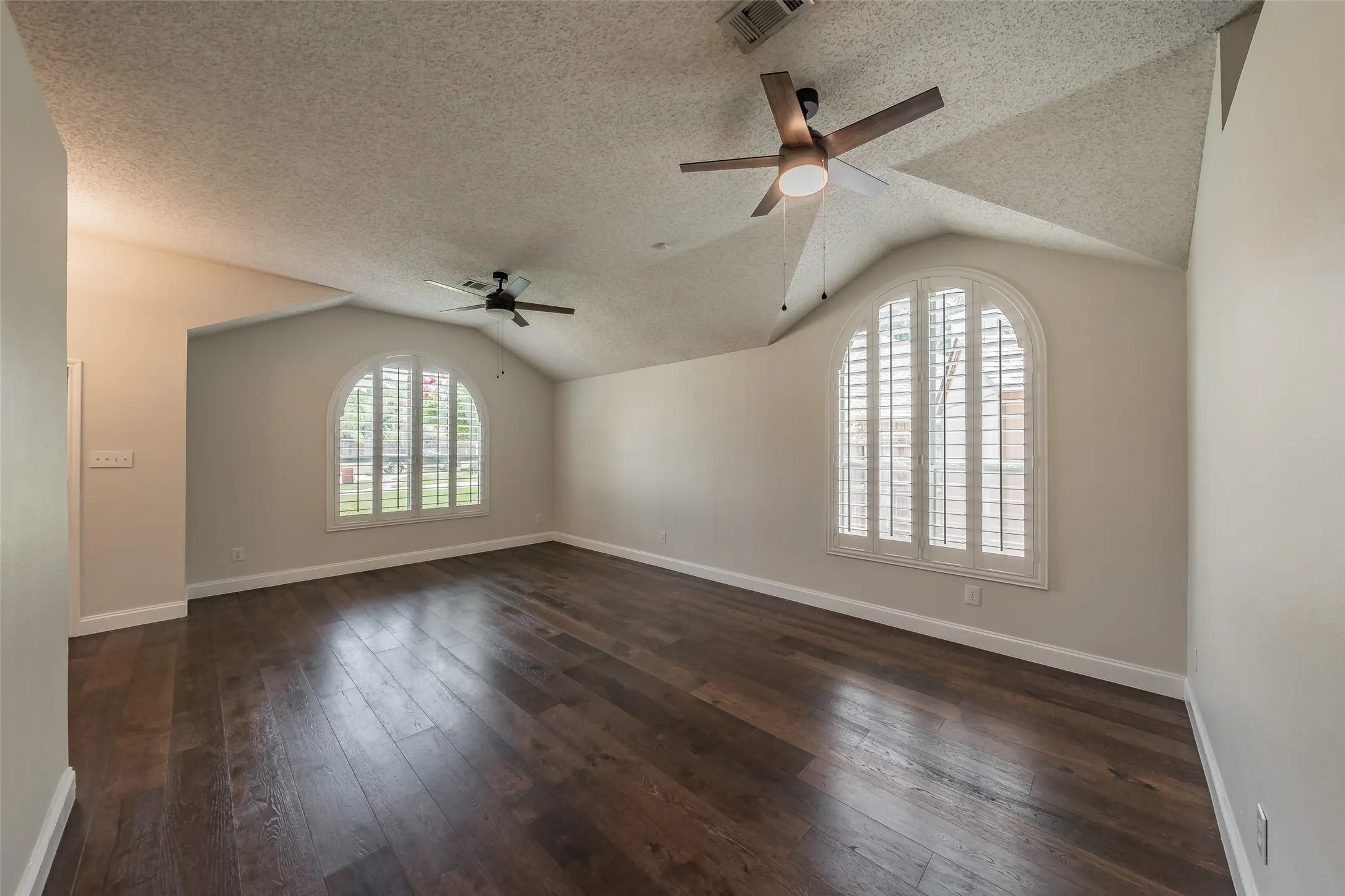 Spacious room featuring vaulted ceilings, rich wood-finish flooring, and two arched windows with plantation shutters