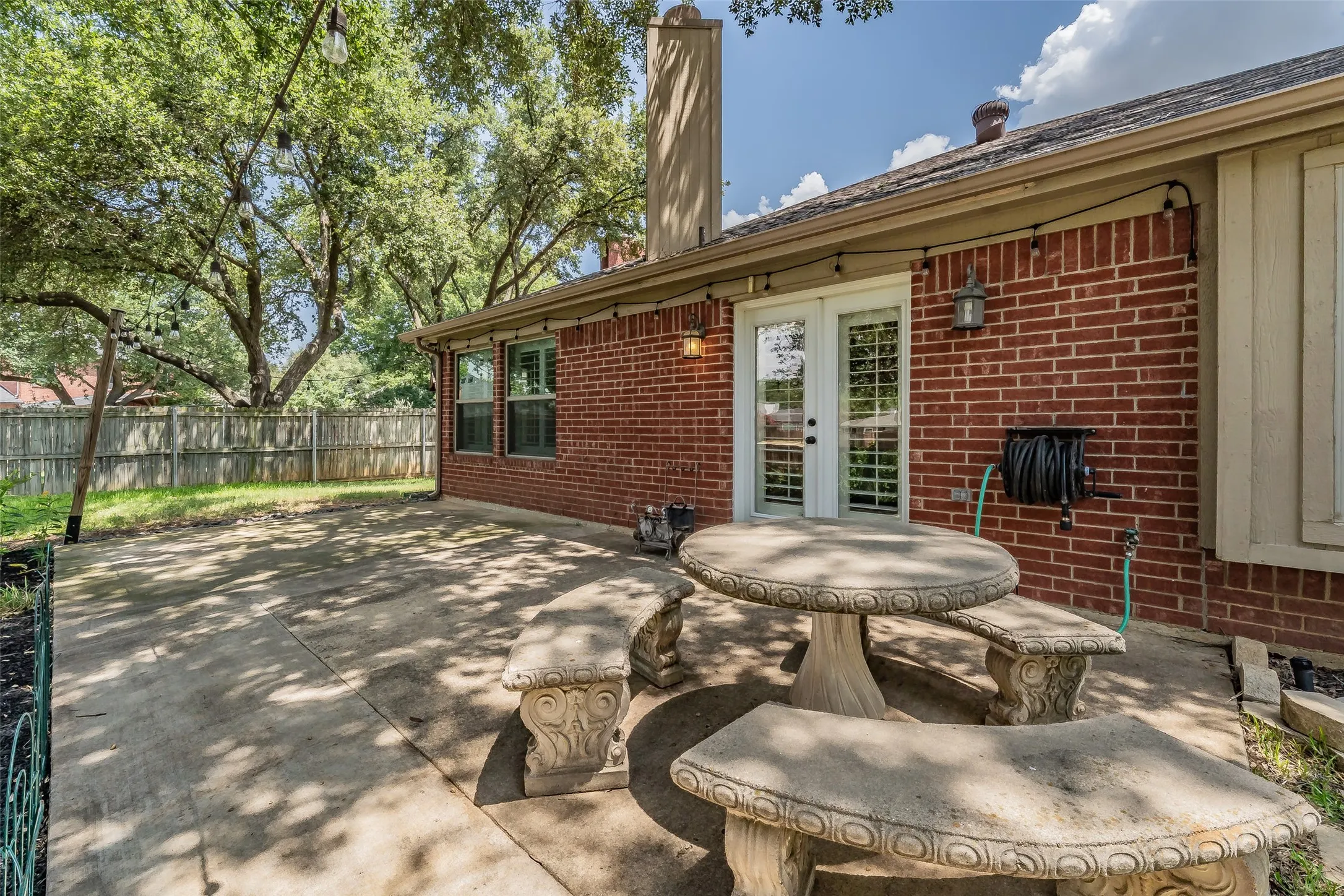 Concrete patio area featuring a red brick exterior, double glass panel doors with interior shutters, two exterior wall sconces, and a wood privacy fence