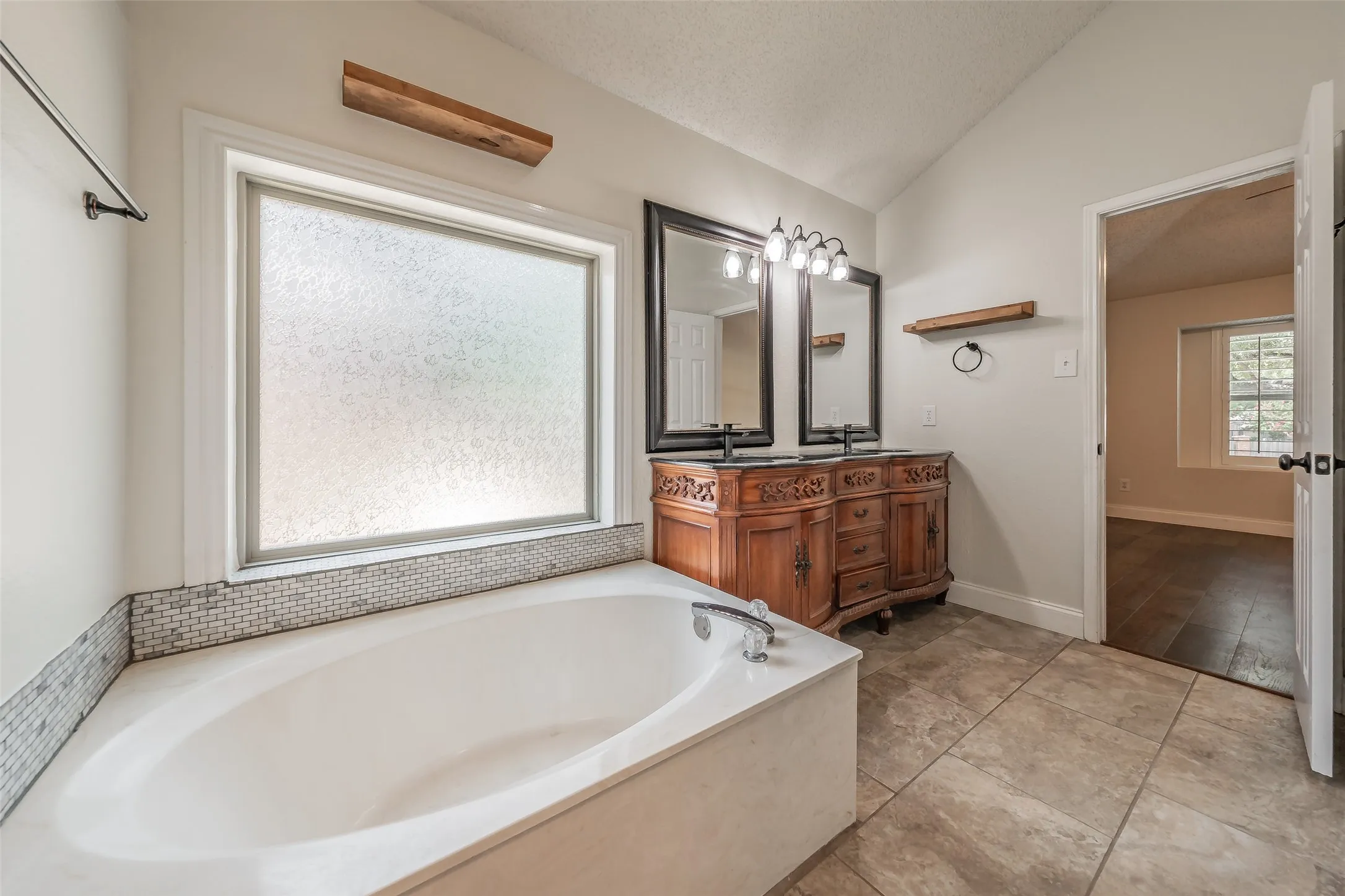 Soaking tub with a tiled surround, dual basin vanity with framed mirrors, and large format floor tiling