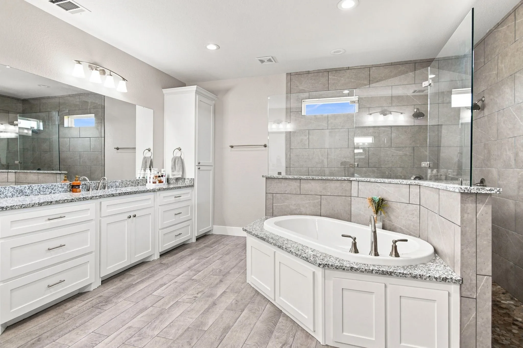 Spacious bathroom featuring a dual vanity with granite countertops, a built-in soaking tub, a walk-in shower with glass enclosure, and wood-finish flooring