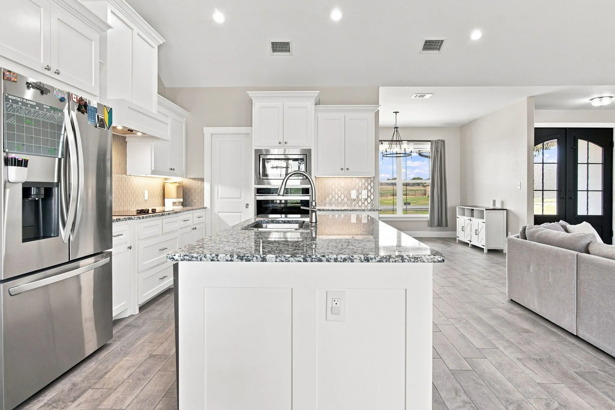 Open-concept kitchen featuring a large central island with a granite countertop, white shaker cabinetry, stainless steel appliances, and wood-finish flooring
