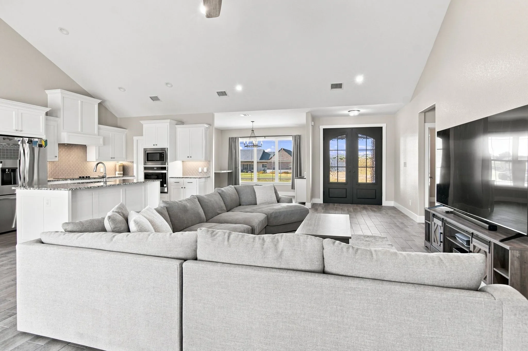 Spacious open-concept living area featuring vaulted ceilings, wood-finish flooring, and a kitchen with white cabinetry, stainless steel appliances, and a tile backsplash