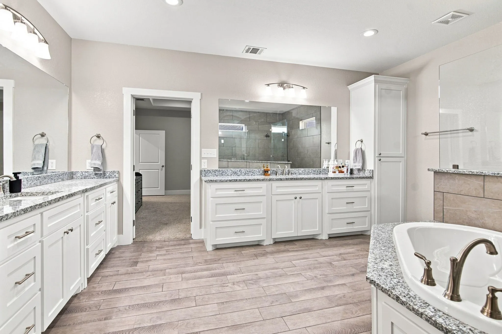 Spacious bathroom featuring wood-finish flooring and dual vanity areas with white cabinetry and speckled countertops