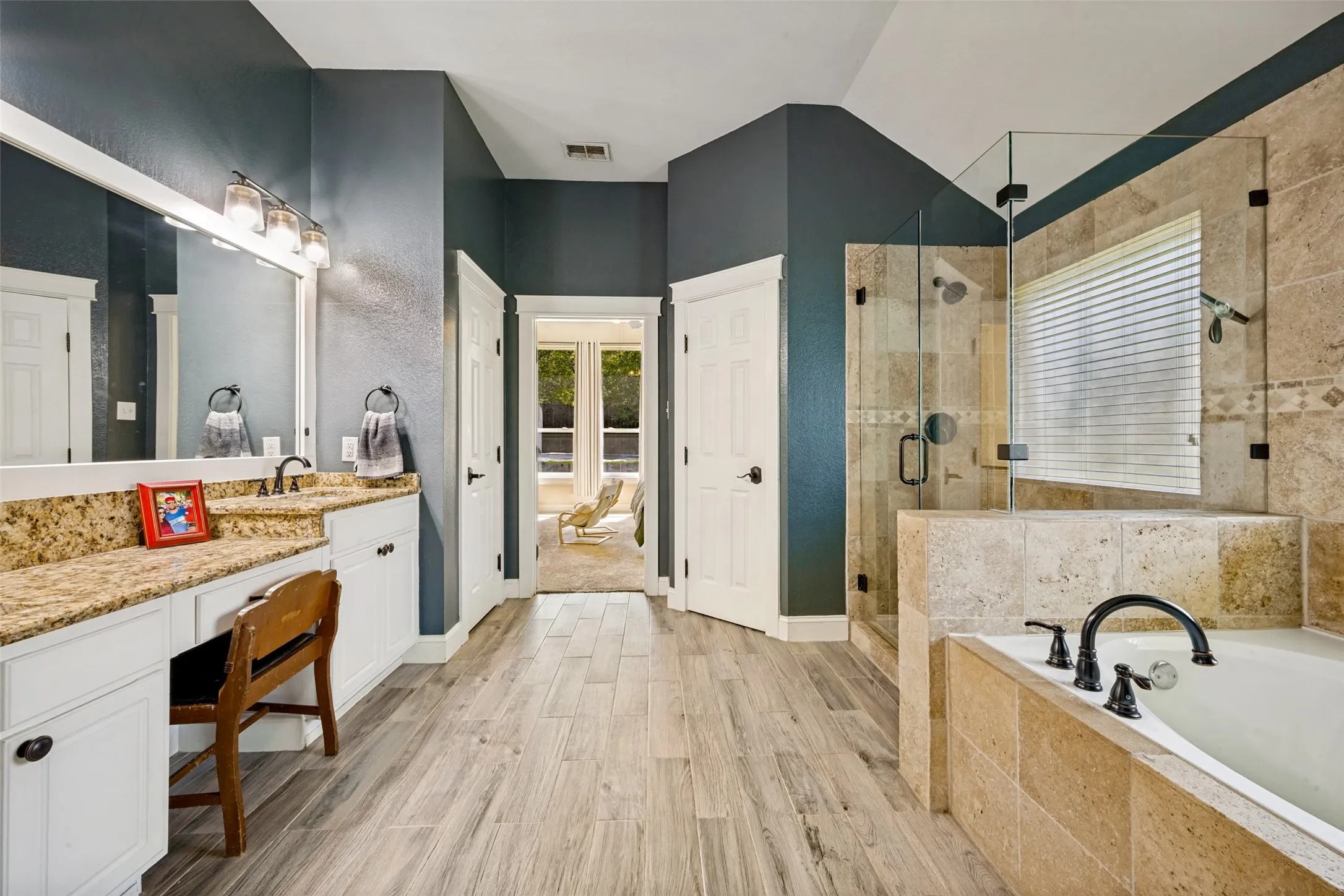 Spacious bathroom featuring a double vanity with granite countertops, a built-in vanity desk, a walk-in shower with glass enclosure, a built-in soaking tub, and wood-finish flooring