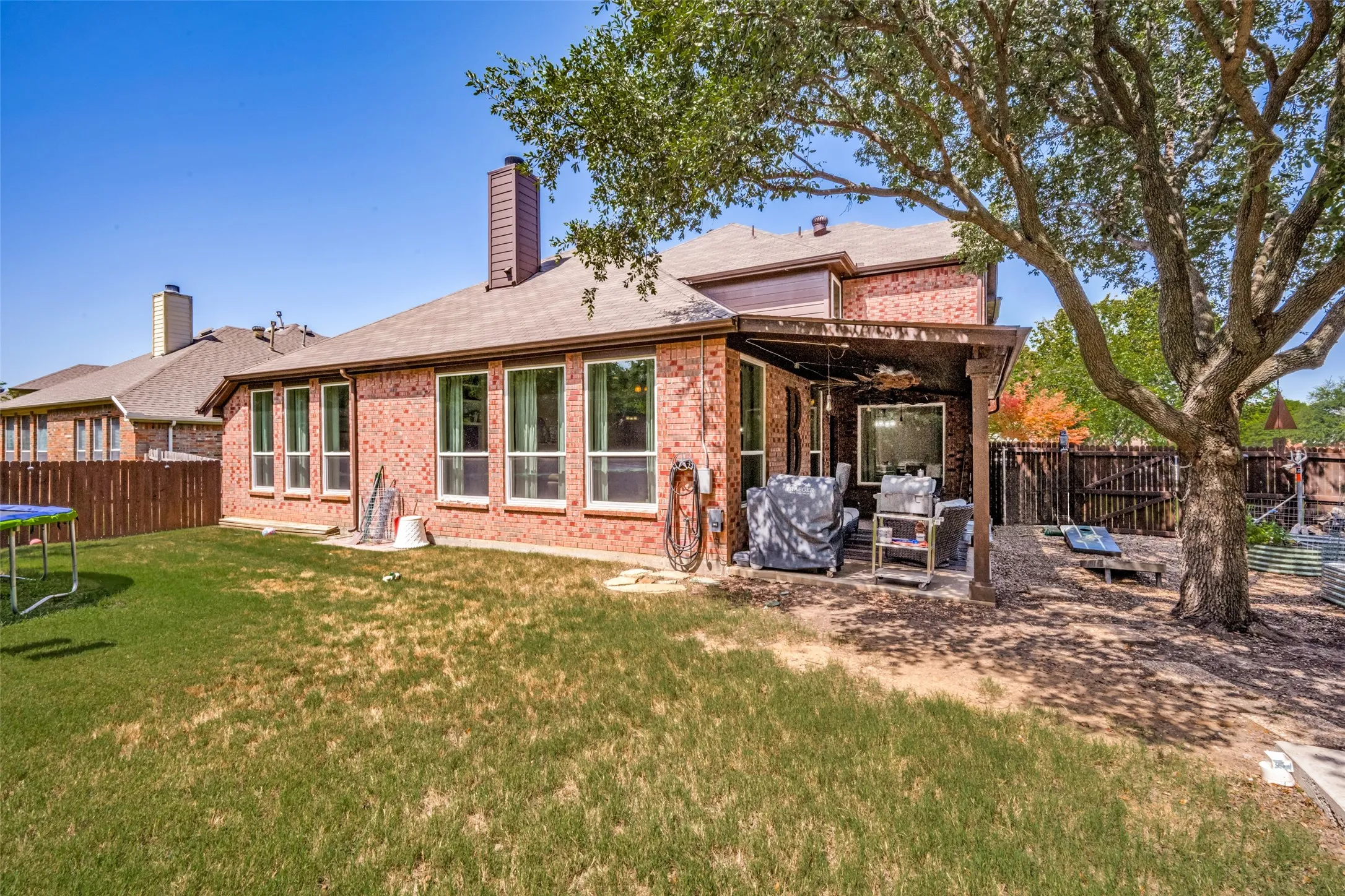 Expansive backyard featuring a covered patio, brick exterior, multiple windows, a mature tree, and a wooden fence