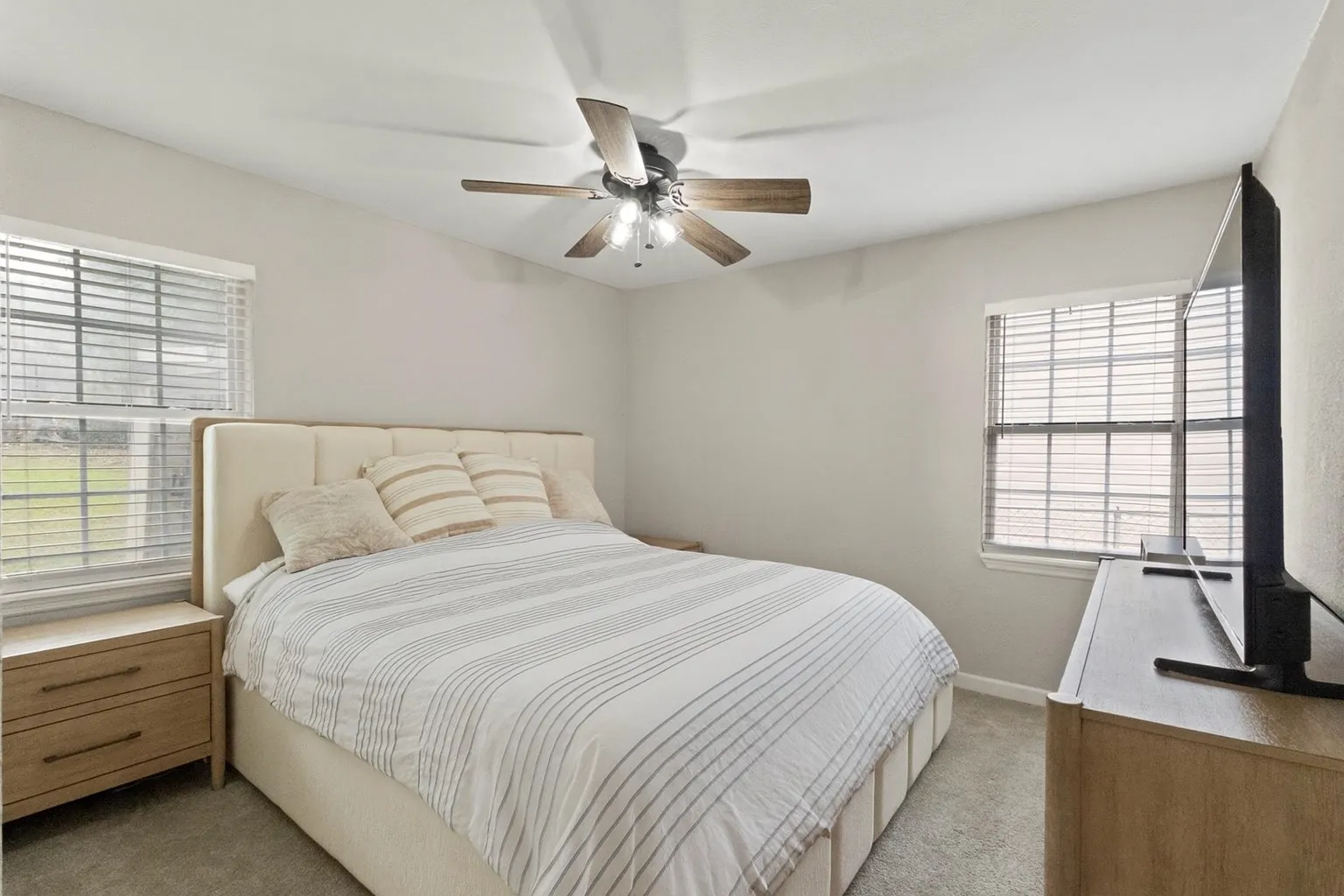 Carpeted bedroom with a ceiling fan light fixture, two windows with blinds, and neutral wall tones
