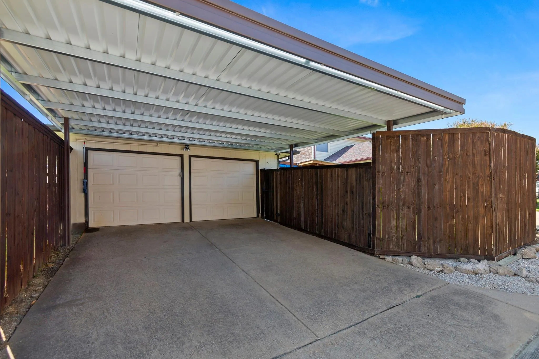 Concrete driveway featuring a metal carport structure with a corrugated roof, two paneled garage doors, and perimeter fencing