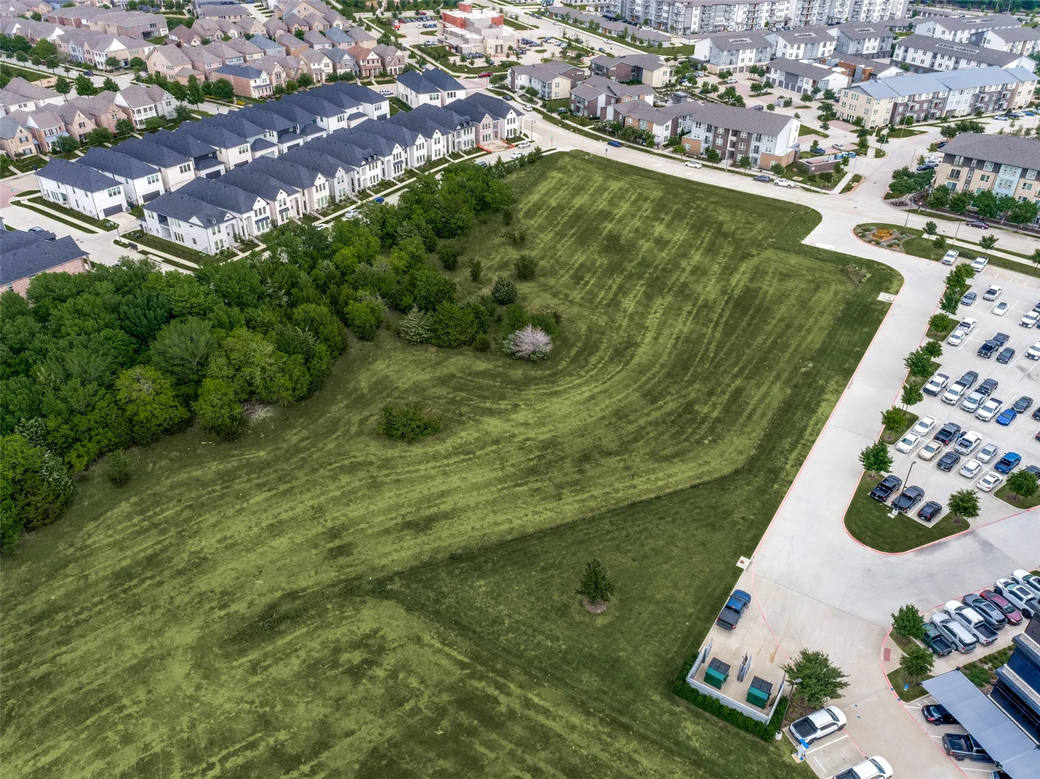 Expansive green space featuring mature trees and manicured lawns, bordered by modern residential developments and commercial parking areas