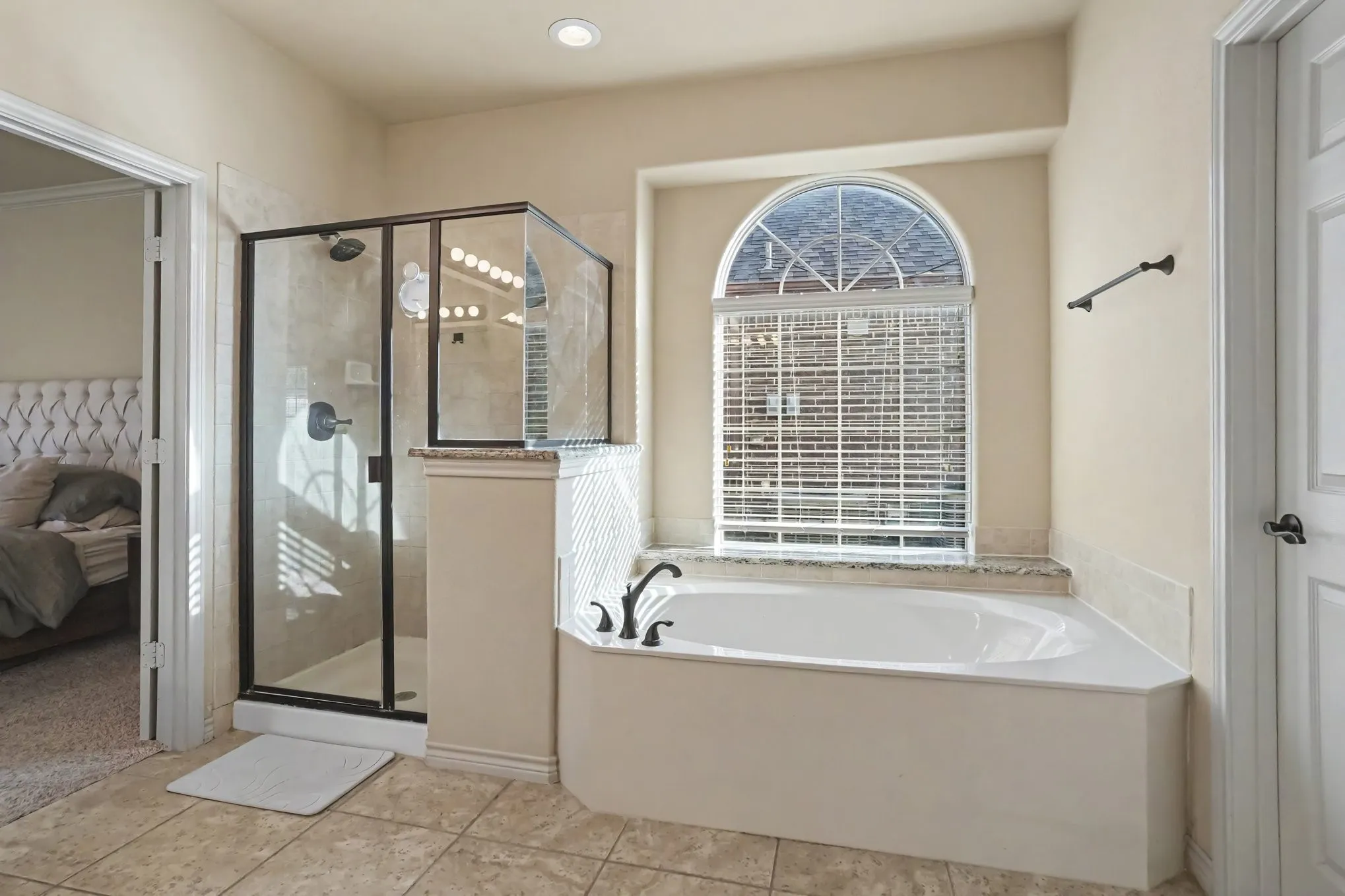 Spacious bathroom featuring a built-in soaking tub with a granite surround, an arched window, a glass-enclosed shower, and tile flooring