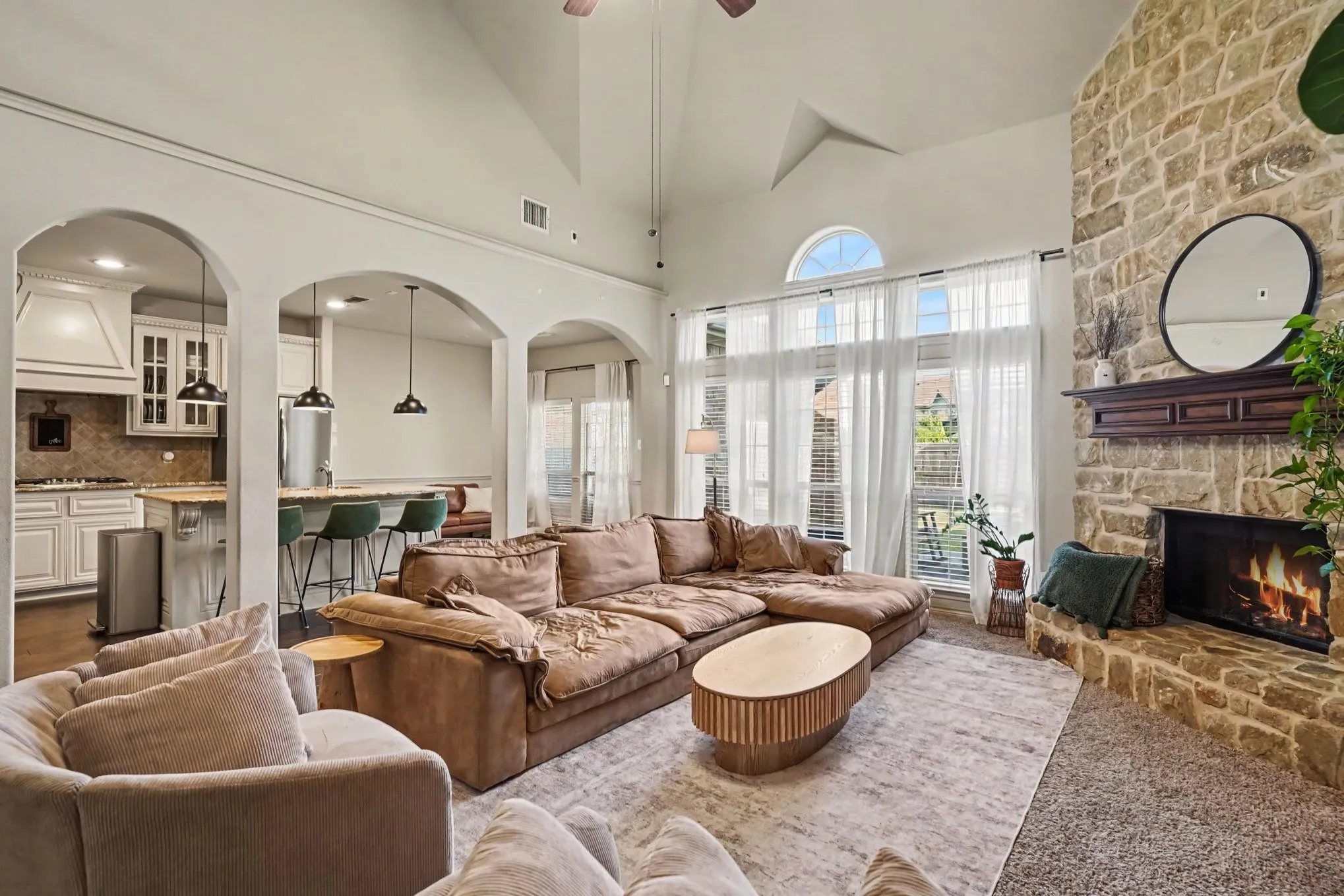 Spacious living area featuring a stone fireplace with a wood mantel, vaulted ceilings, and large windows with an arched transom