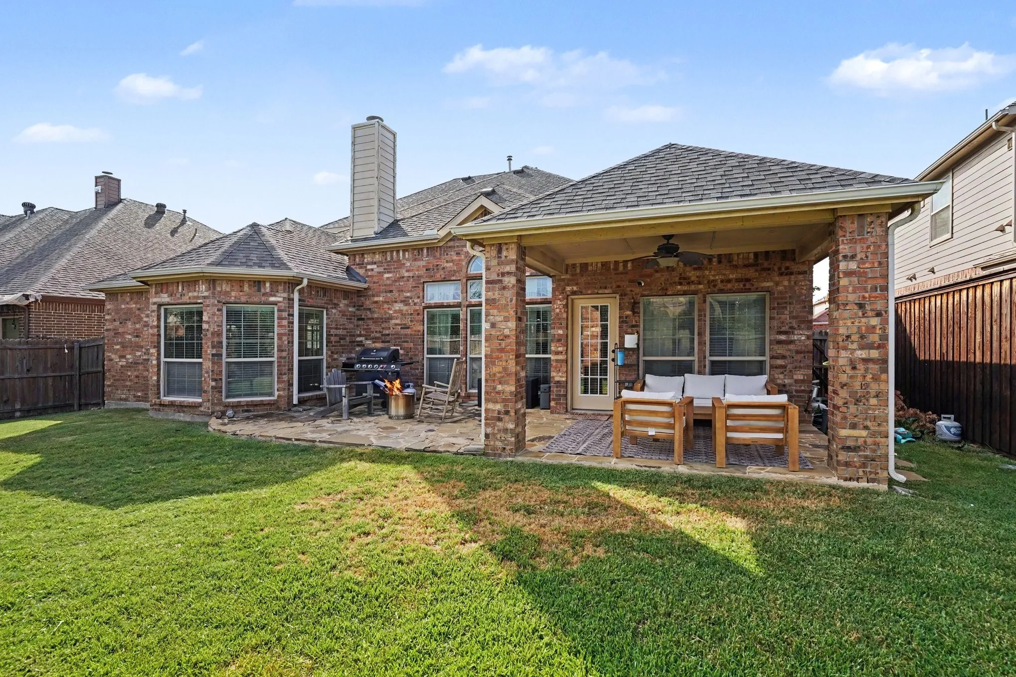 Expansive backyard featuring a covered patio with a ceiling fan, a stone patio area, a brick exterior with multiple windows, and a privacy fence