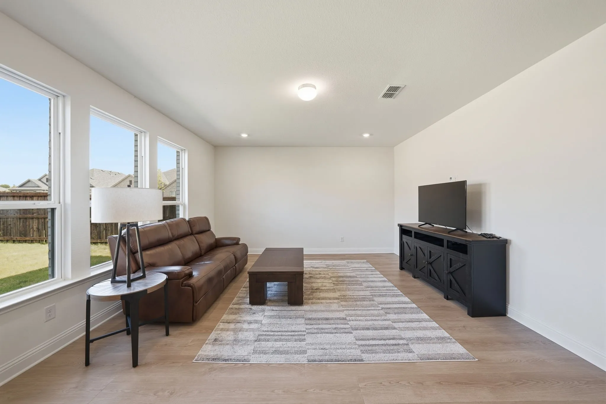Spacious living area featuring light wood-finish flooring, multiple large windows providing abundant natural light, recessed ceiling lighting, and a neutral color palette