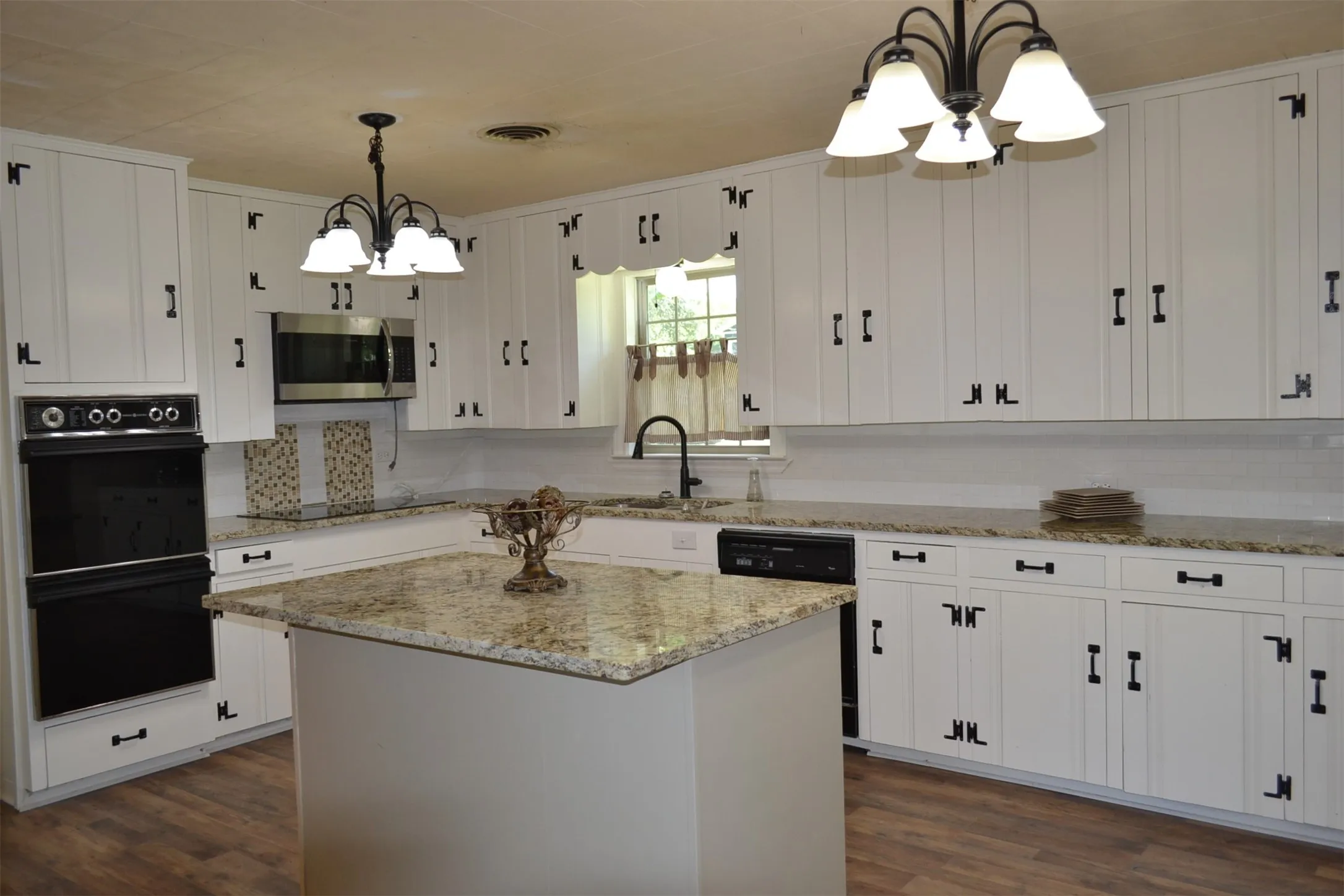 Kitchen featuring white cabinetry with black hardware, double wall oven, microwave, an island with granite countertops, and wood-finish flooring