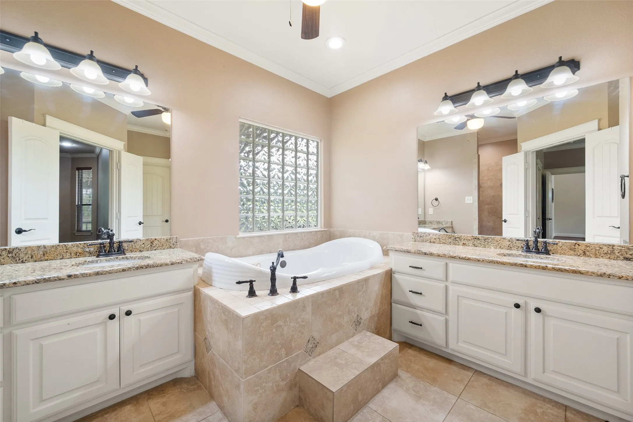 Primary bathroom with dual granite vanities, jetted soaking tub, built-in cabinetry and decorative glass-block window.