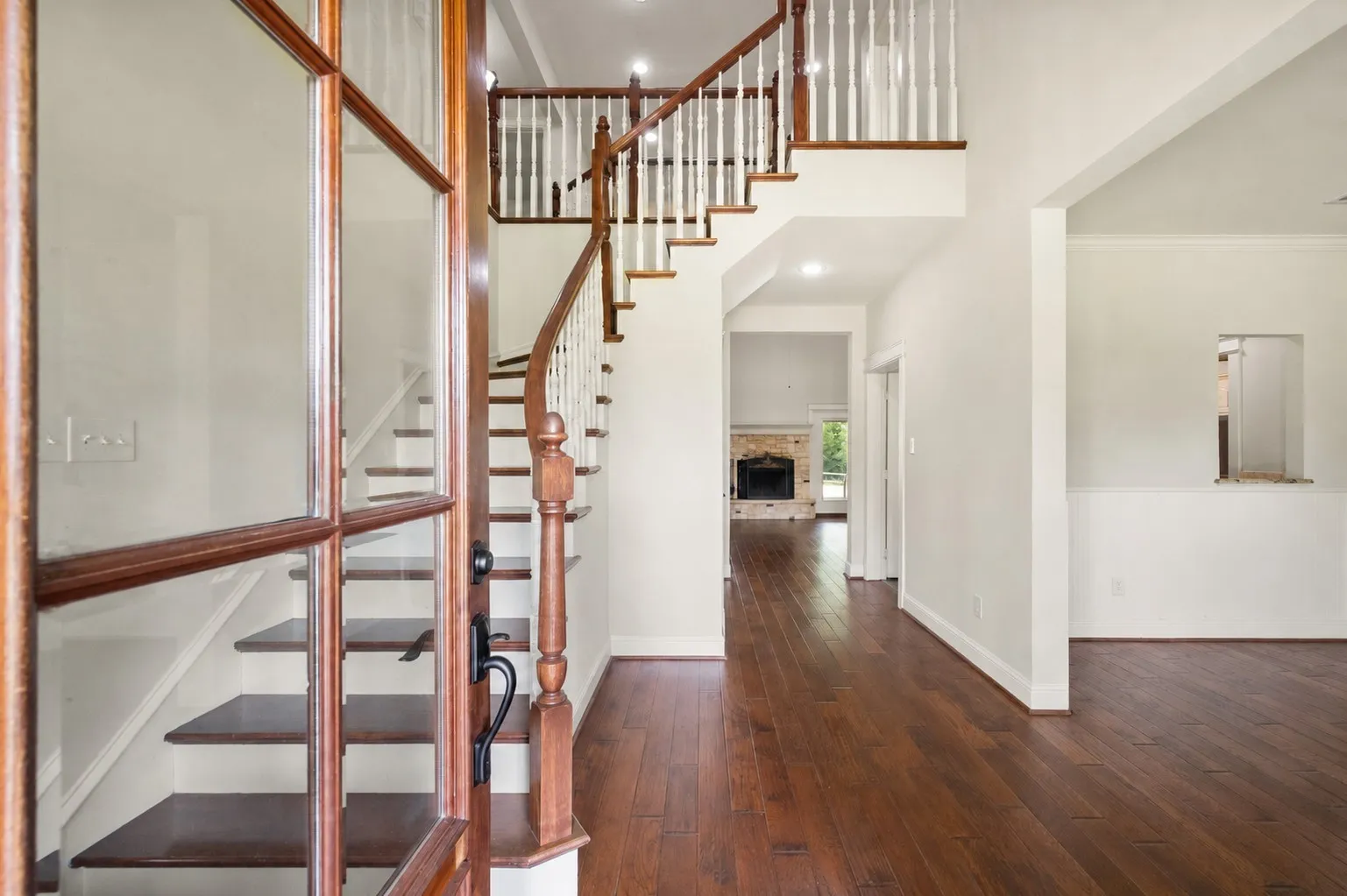 Welcoming two-story foyer with hardwood flooring, natural wood staircase and views toward the stone fireplace.