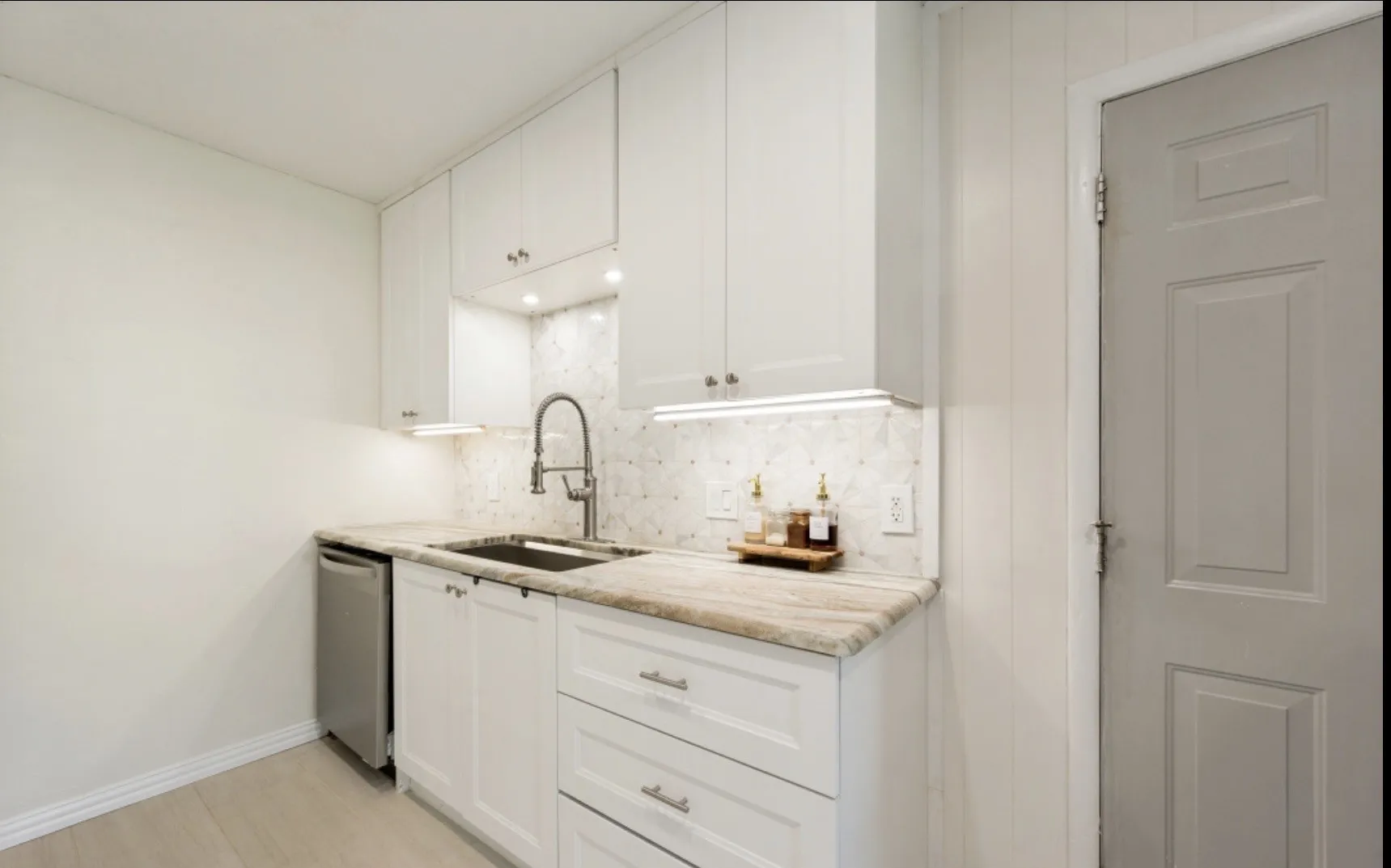 Kitchenette featuring white cabinetry with brushed nickel hardware, stone countertop, stainless steel sink with gooseneck faucet, tiled backsplash, and under-cabinet lighting