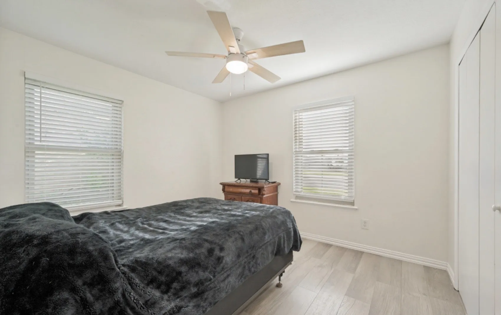 Bedroom featuring light wood-finish flooring, two window openings with blinds, a ceiling fan with light fixture, and a built-in closet with bi-fold doors