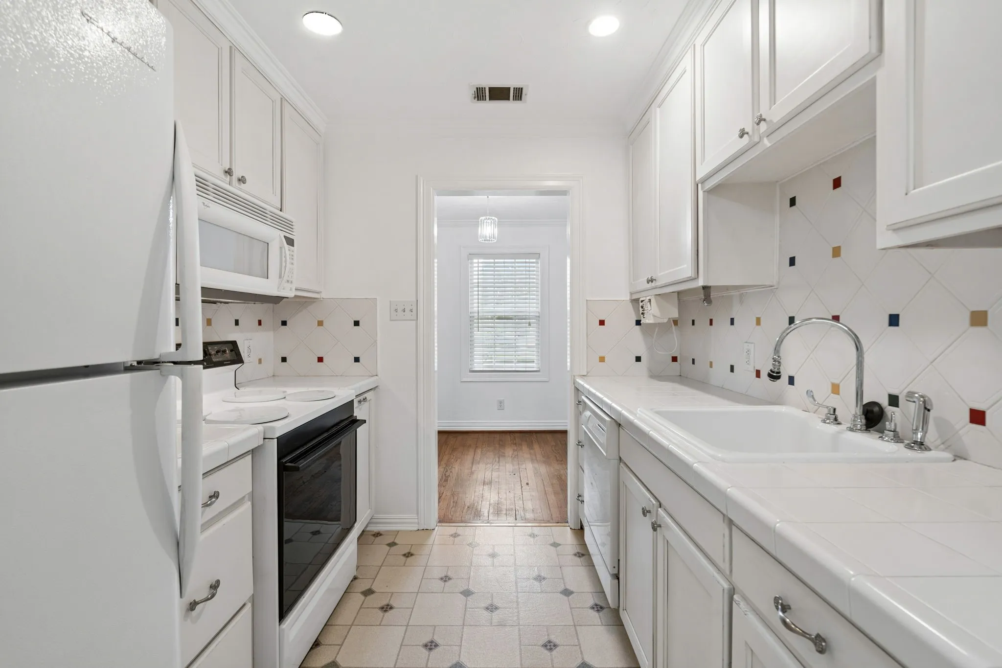 Galley kitchen featuring white cabinetry, integrated white appliances, and a tile backsplash with colorful accents