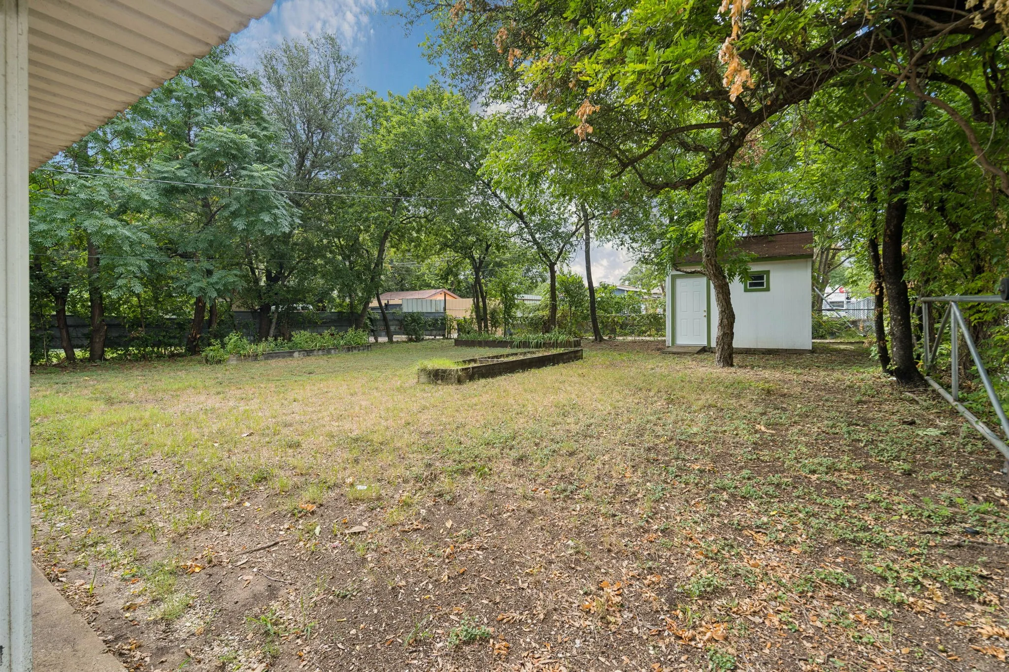 Expansive backyard featuring mature trees, a white storage shed with a brown roof, and raised garden beds