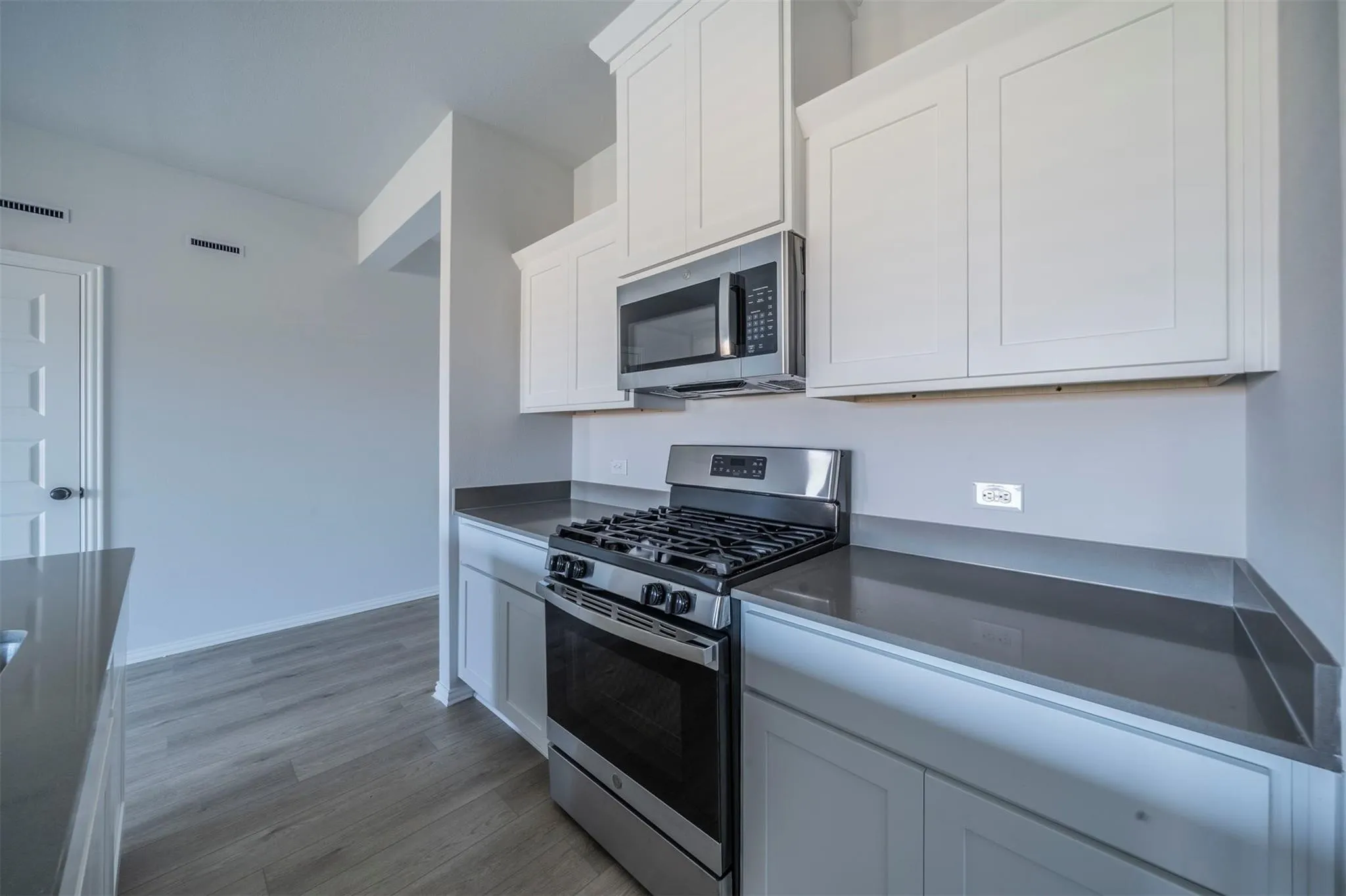 Kitchen featuring white shaker-style cabinetry, stainless steel oven with gas range, stainless steel microwave, dark gray countertops, and wood-finish flooring