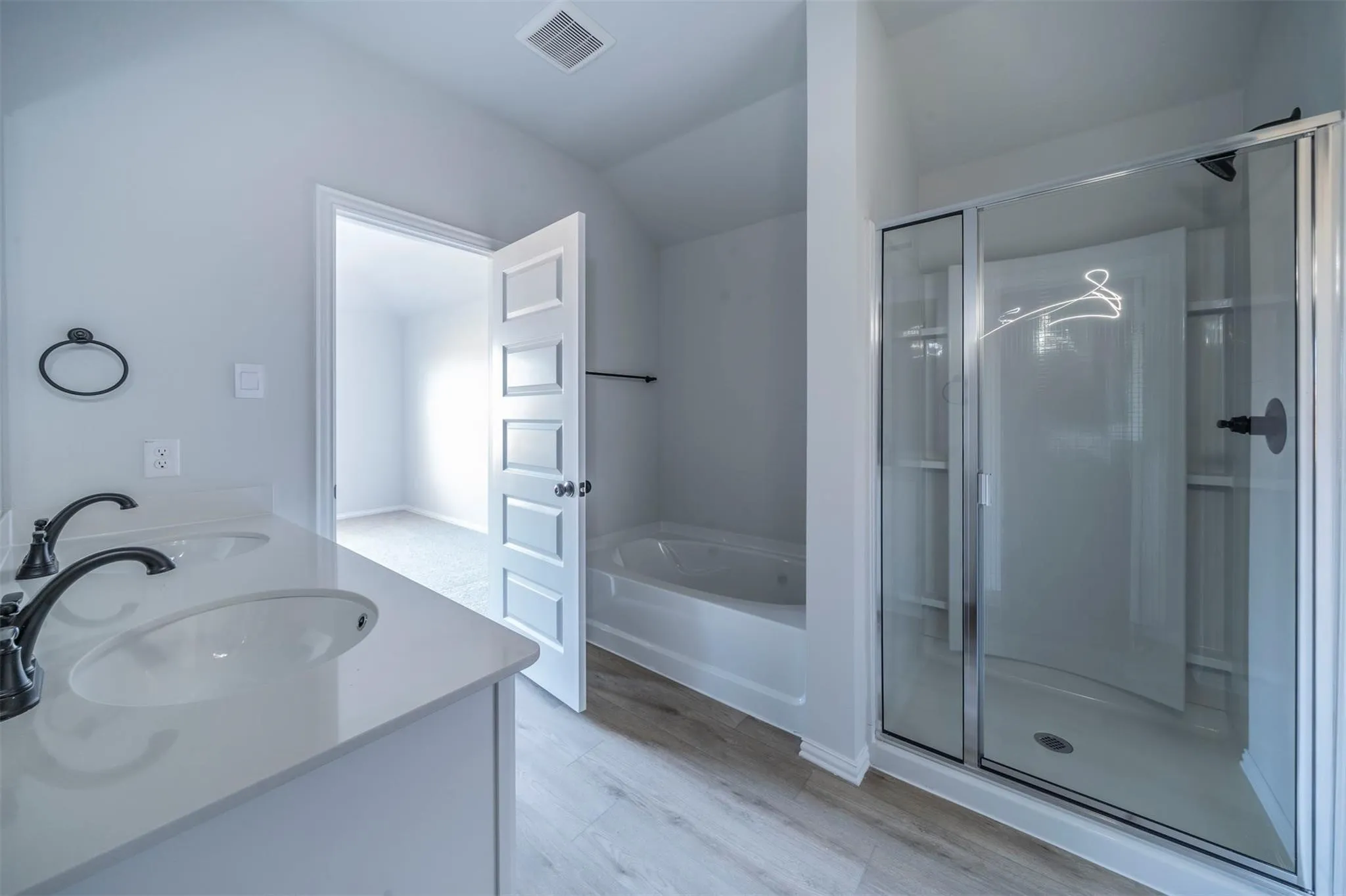 Modern bathroom featuring a dual vanity with white countertops and matte black fixtures, a built-in tub, a glass-enclosed shower, and light wood-finish flooring