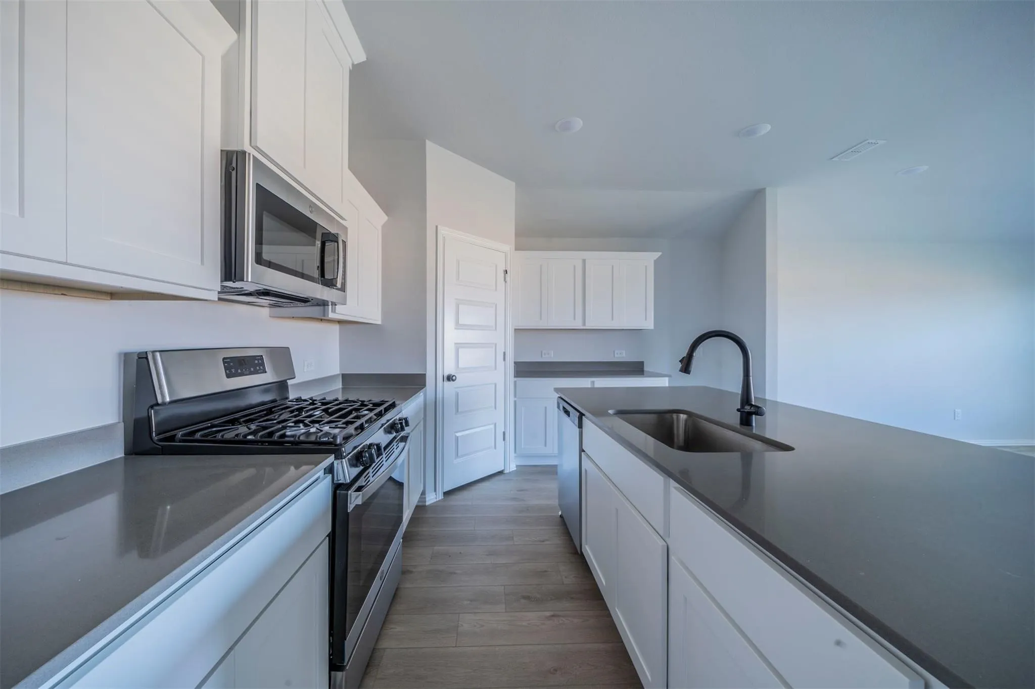 Bright kitchen featuring white shaker cabinetry, stainless steel appliances, dark countertops, and a central island with an undermount sink and matte black faucet