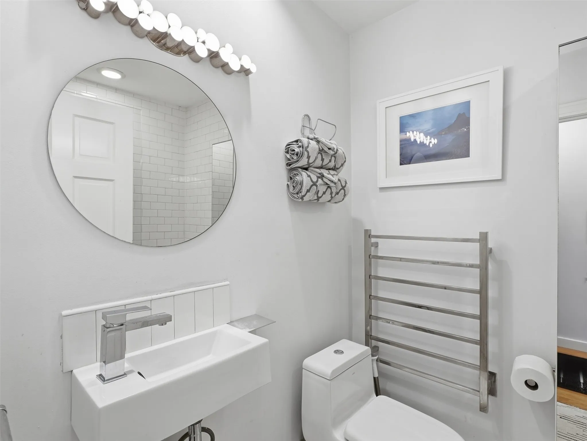 Contemporary bathroom featuring a wall-mounted rectangular sink with a single-handle chrome faucet, a circular mirror, a decorative vanity light fixture, a toilet, and a wall-mounted towel warmer