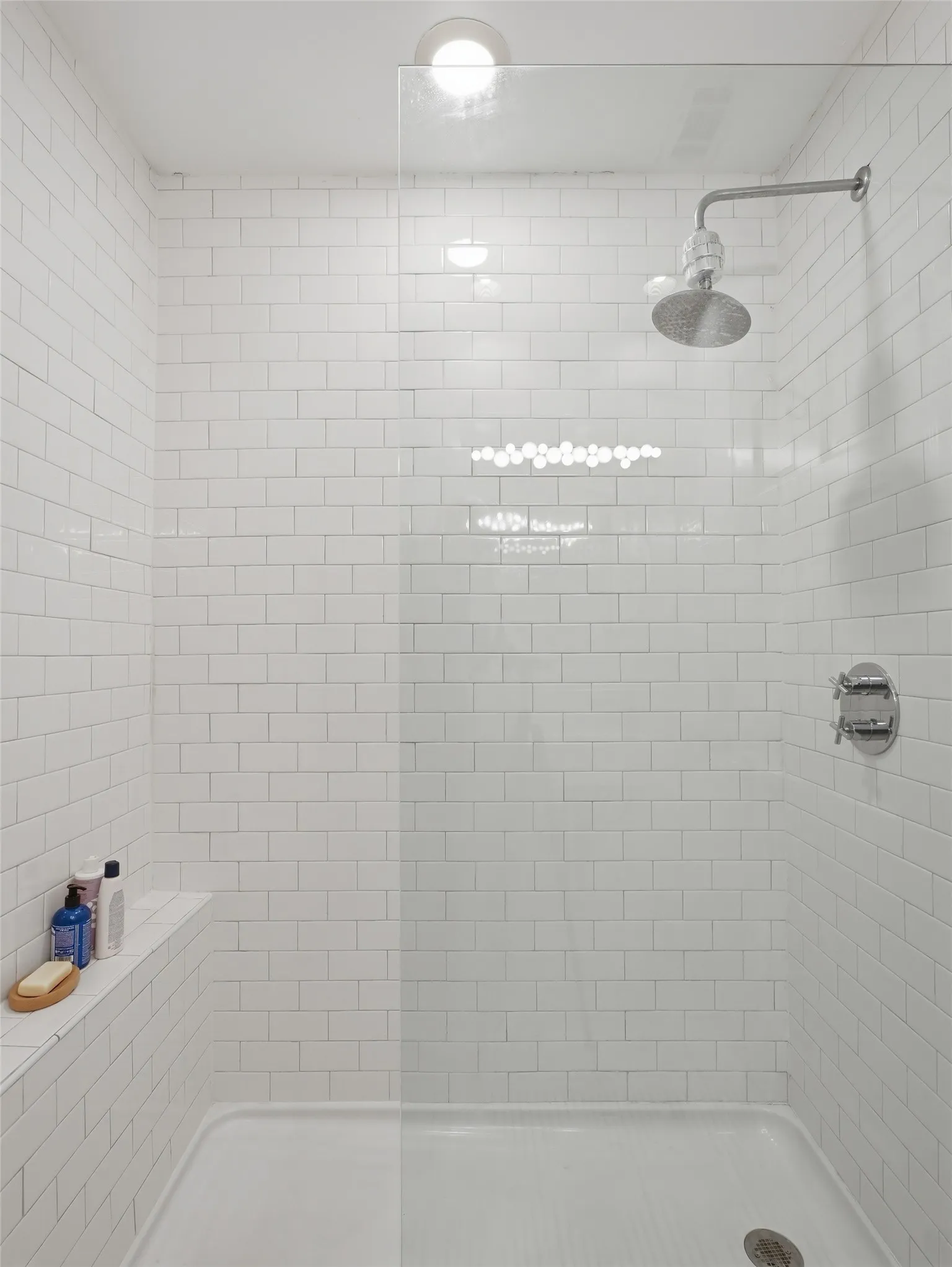 Tiled shower enclosure featuring white subway tile walls, a built-in shelving niche, a clear glass panel, and a wall-mounted showerhead with control fixtures