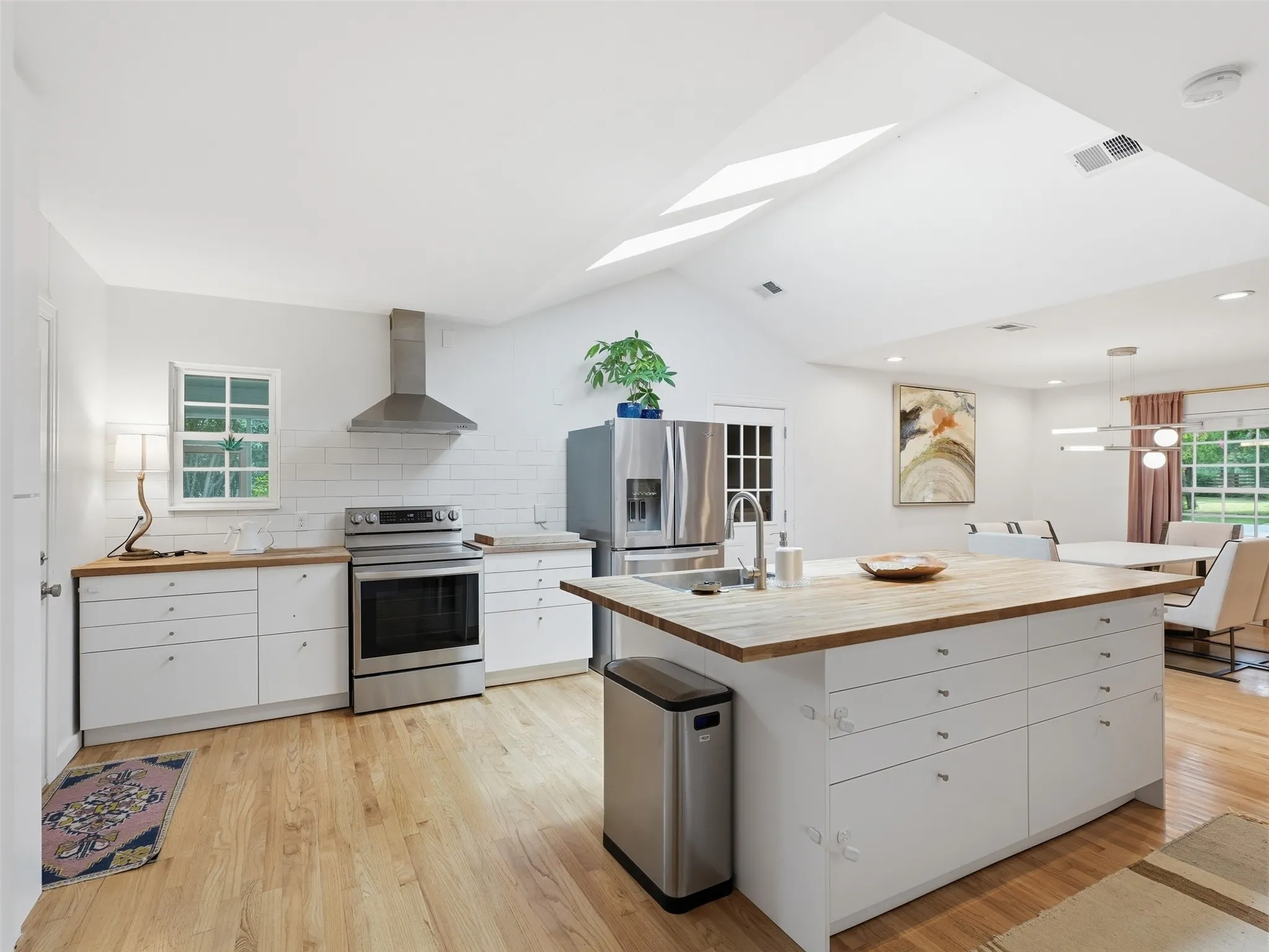 Vaulted ceilings with skylights illuminate this kitchen featuring stainless steel appliances, a center island with a wood-finish countertop, white cabinetry with silver hardware, and wood-finish flooring