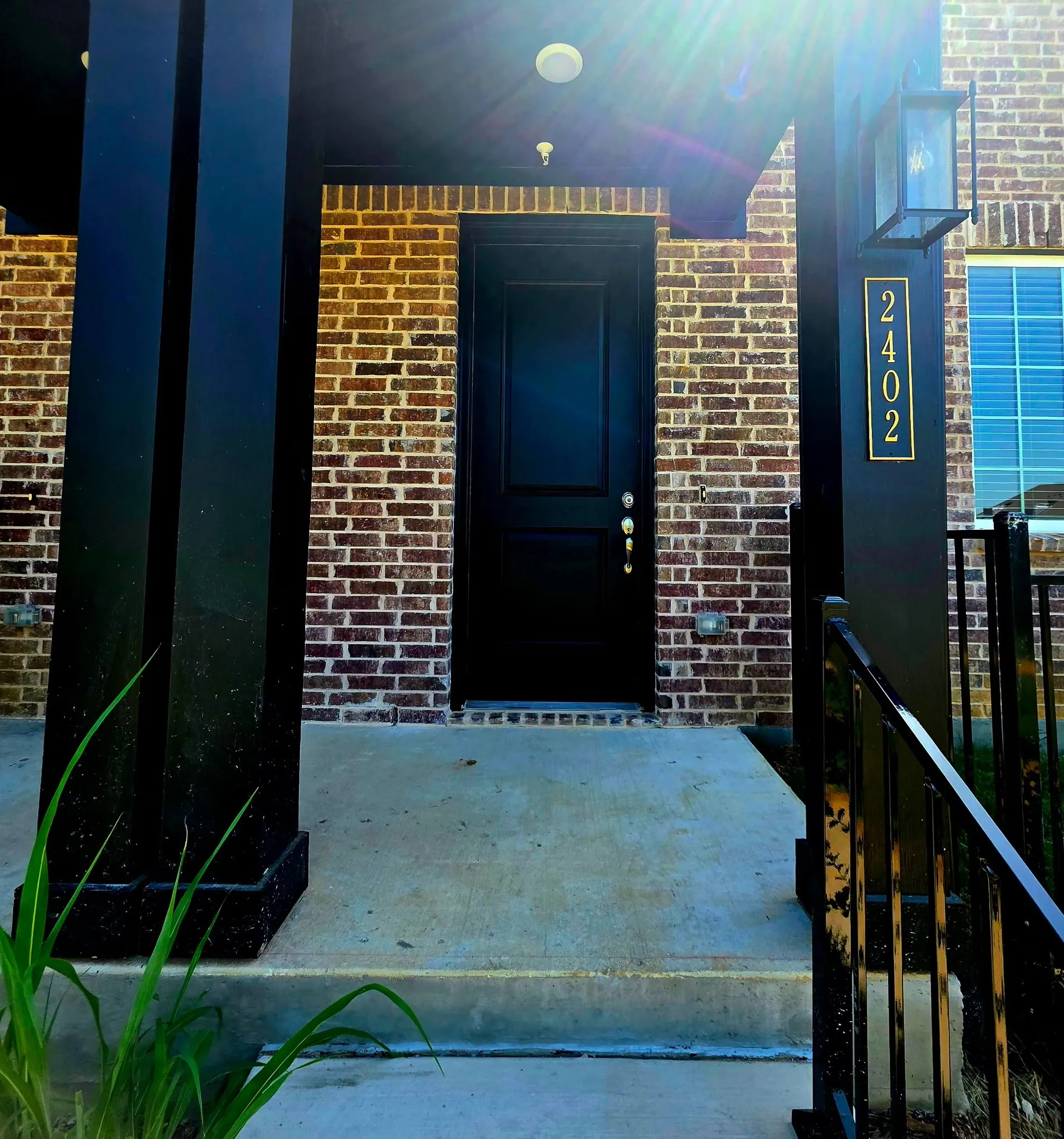 Brick exterior with a prominent black entry door, featuring a concrete walkway, black columns, and a black wrought iron railing