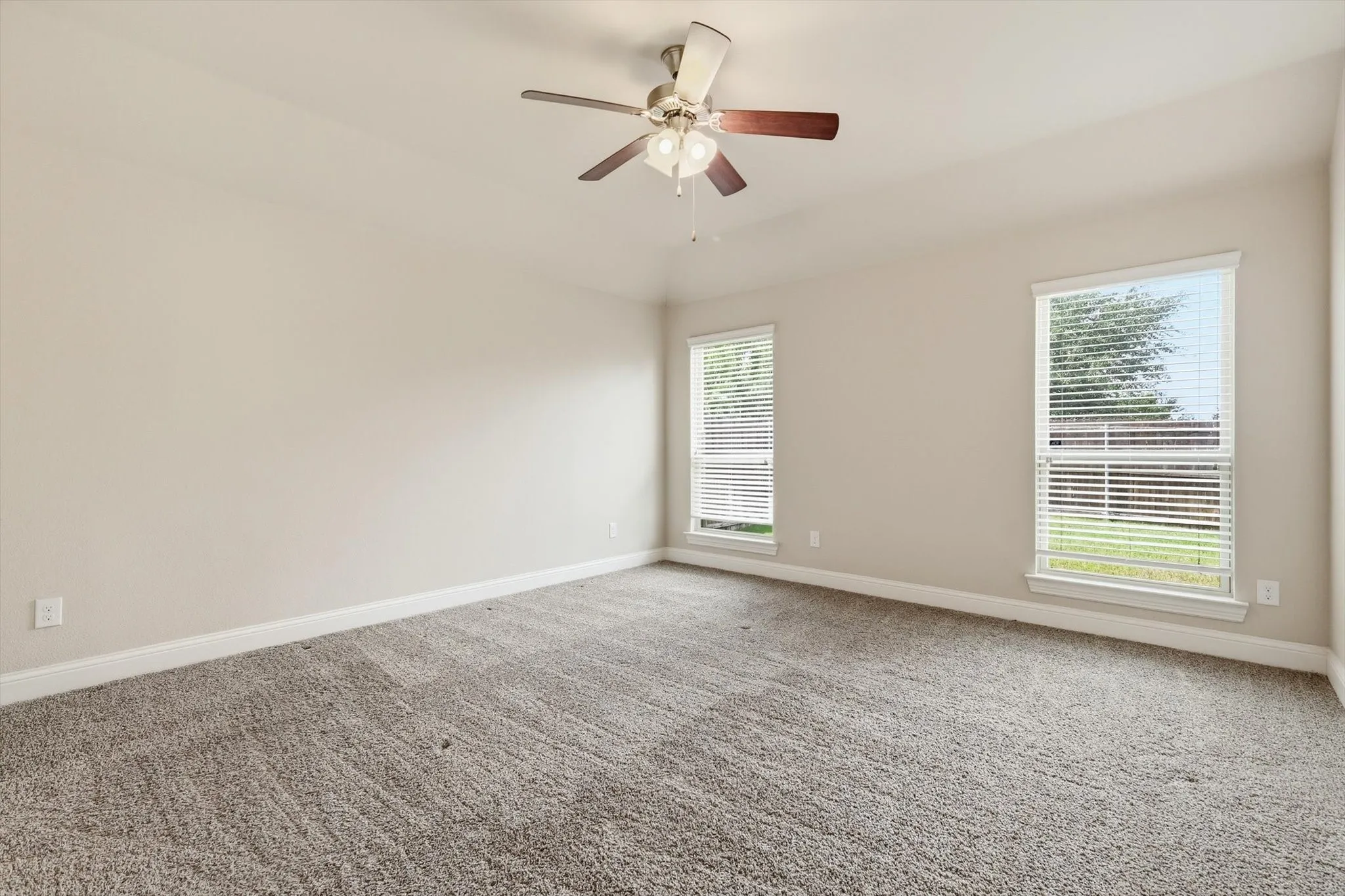 Spacious interior room featuring neutral wall tones, plush carpet flooring, two large windows with blinds, and a ceiling fan with integrated lighting