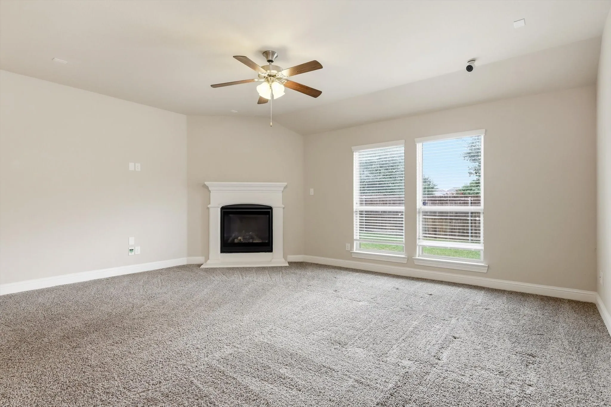 Spacious interior featuring a white mantel fireplace, two large windows with blinds, a ceiling fan, and light gray wall-to-wall carpeting