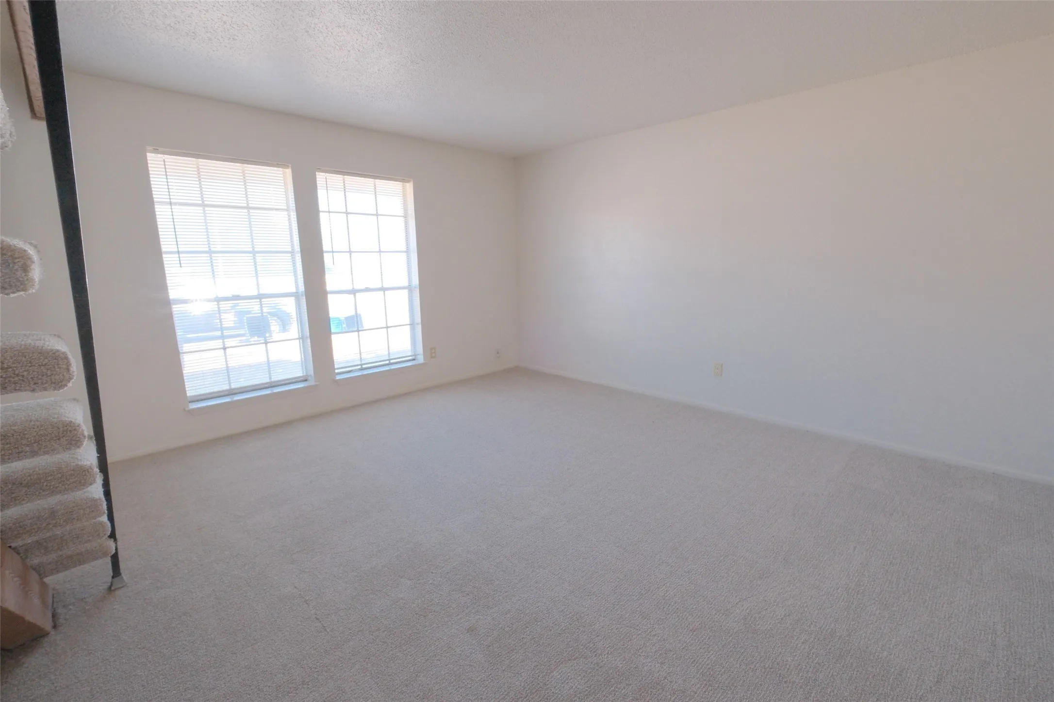 Spacious room featuring neutral carpeting, light-toned walls, and two large windows with horizontal blinds