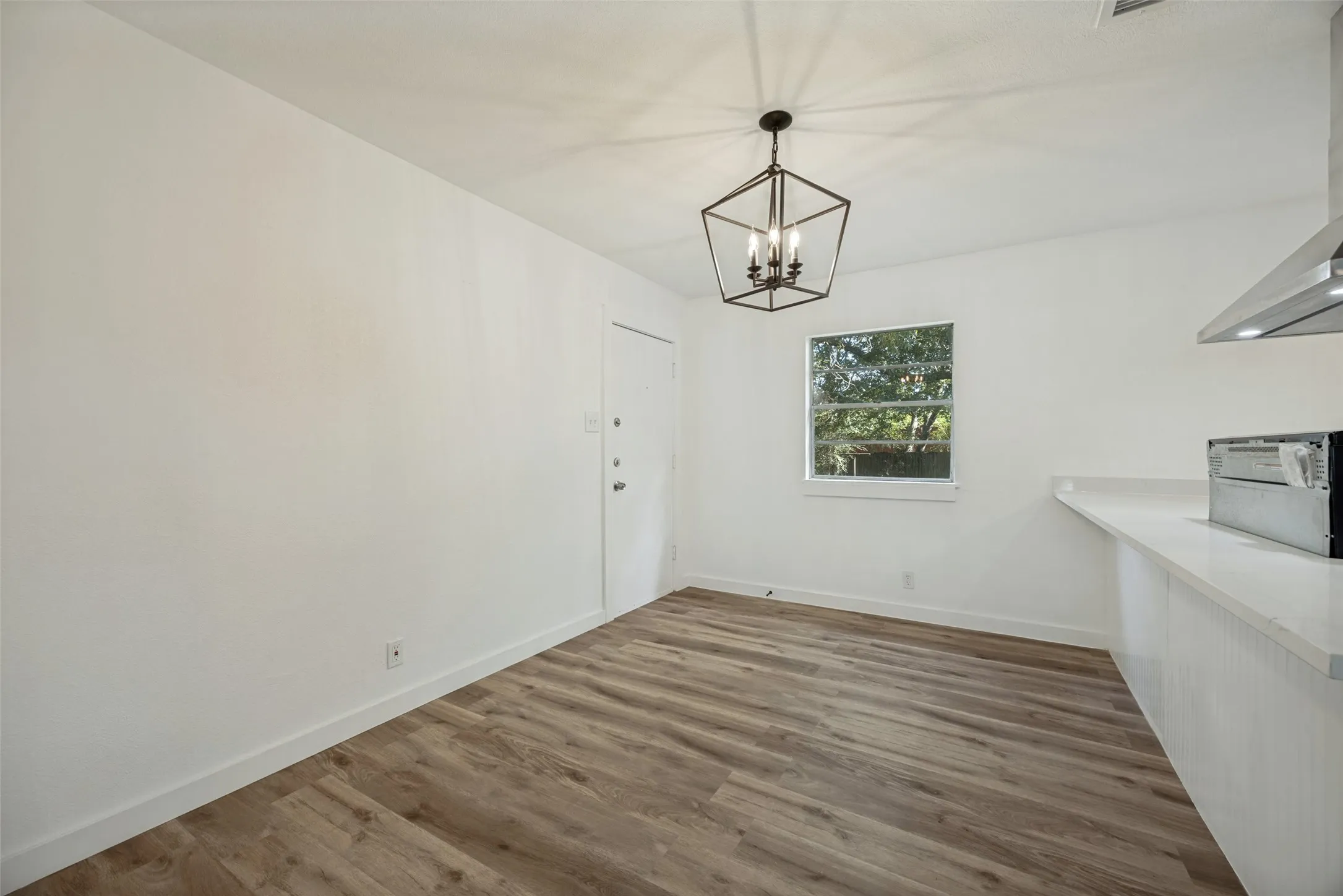 Open-concept dining area featuring wood-finish flooring, a modern geometric chandelier, a window with natural light, a white interior door, and a kitchen peninsula with a light-colored countertop