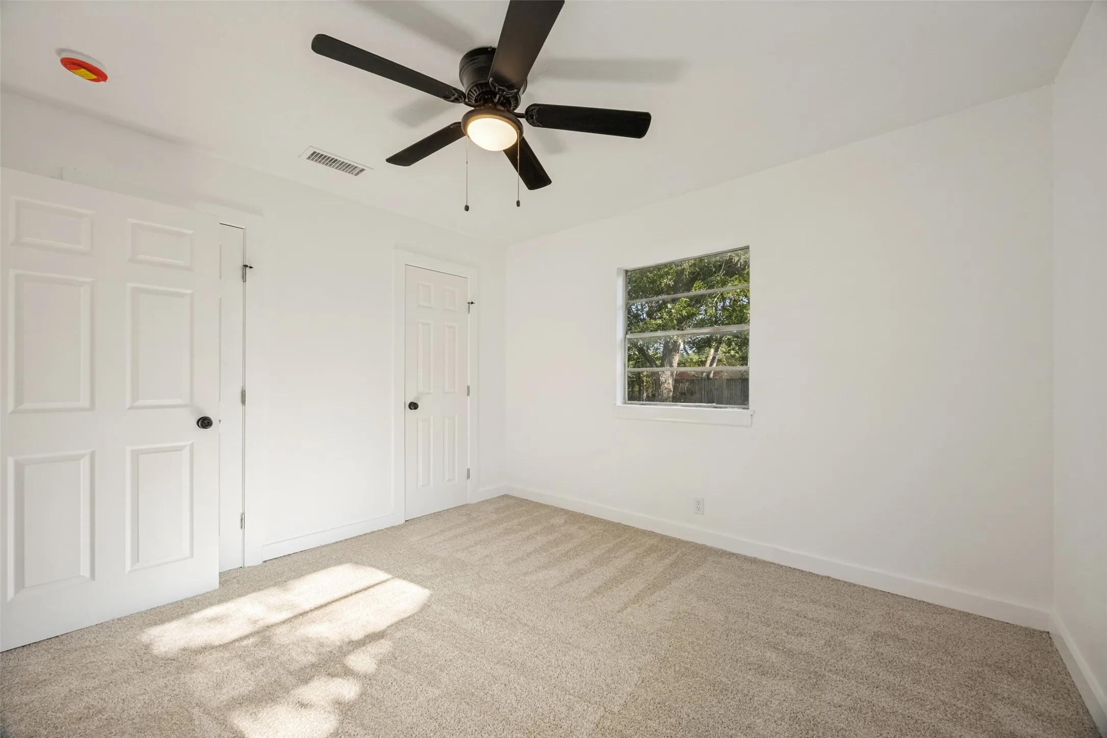 Neutral-toned carpeted room featuring a ceiling fan with integrated lighting, a single window, and white paneled doors with black hardware