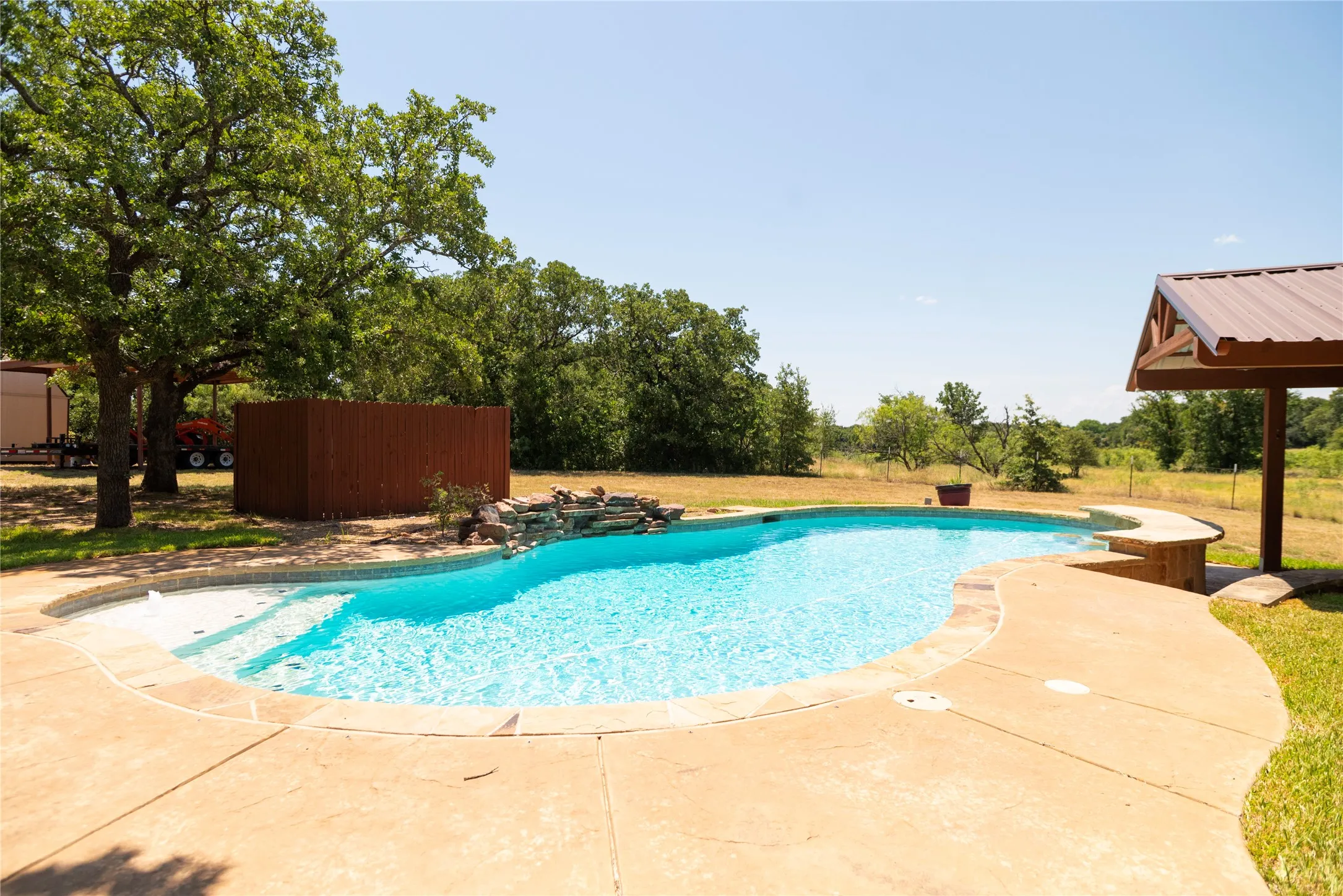 Curved swimming pool featuring a waterfall accent, concrete decking, and a covered patio structure with a metal roof