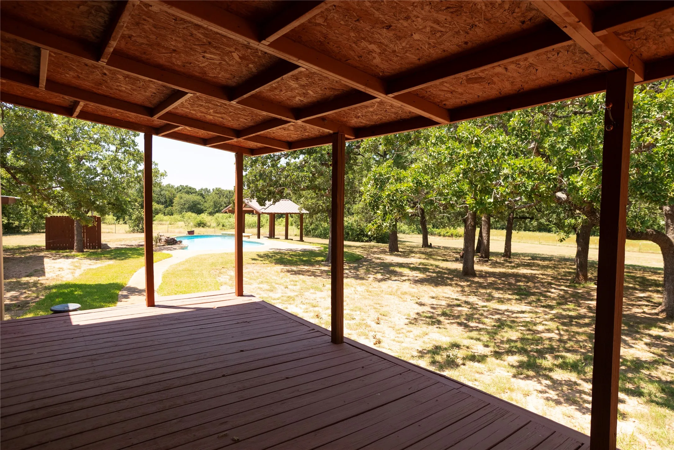 Covered outdoor deck with exposed wood rafters, overlooking a swimming pool, adjacent gazebo, and mature trees