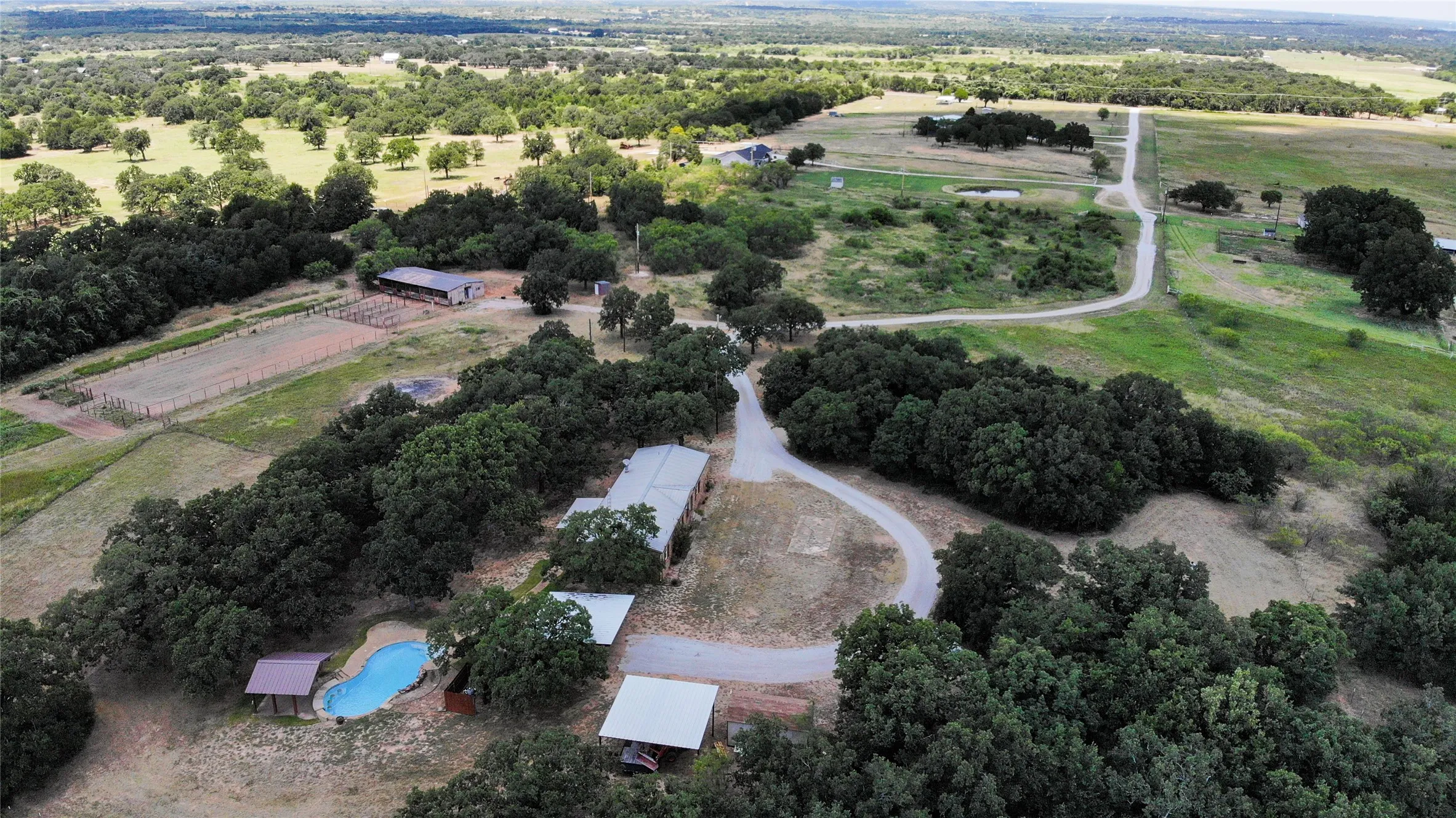 Expansive property featuring a winding driveway, several outbuildings, a blue-tiled swimming pool with a covered patio, and a fenced equestrian arena
